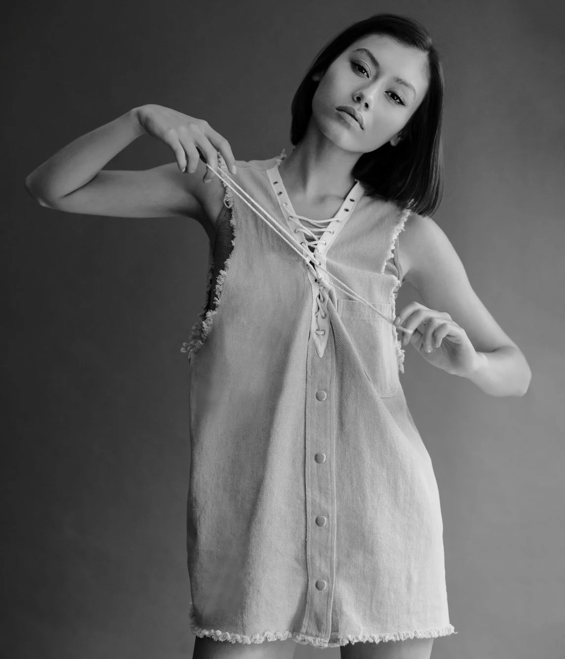 Black and white photo of a young woman with shoulder-length dark hair wearing a sleeveless, distressed denim dress with lace-up front. She is holding a string in both hands, pulling it apart, with a neutral facial expression.