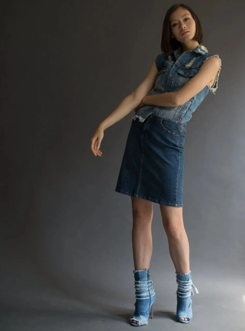 A young woman posing against a gray background, wearing a sleeveless denim vest, a denim skirt, and distressed denim high-heeled shoes.