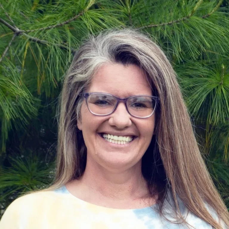 A woman with long, grayish-brown hair and glasses smiling outdoors in front of green pine tree branches.