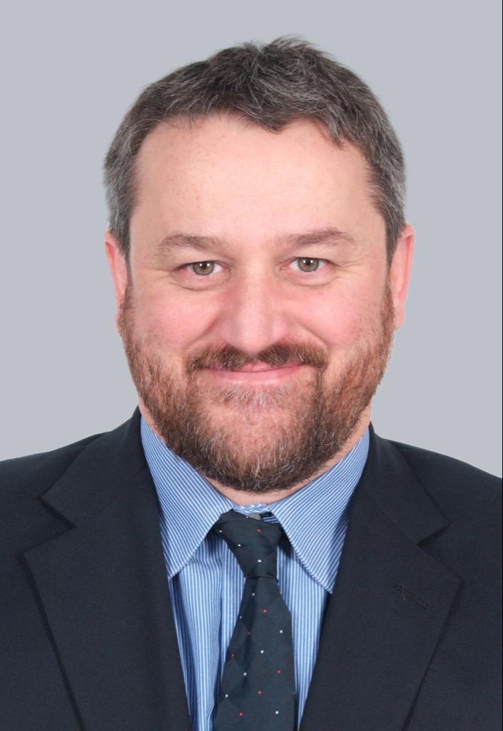 Professional headshot of a smiling man with short, dark hair and a beard, wearing a suit, dress shirt, and tie against a plain gray background.