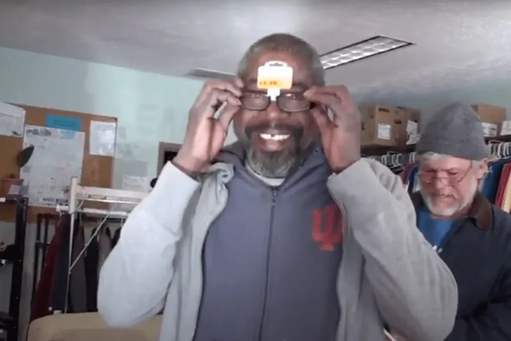 A man smiling and holding glasses on his face in a room with two bulletin boards and clothing racks in the background.