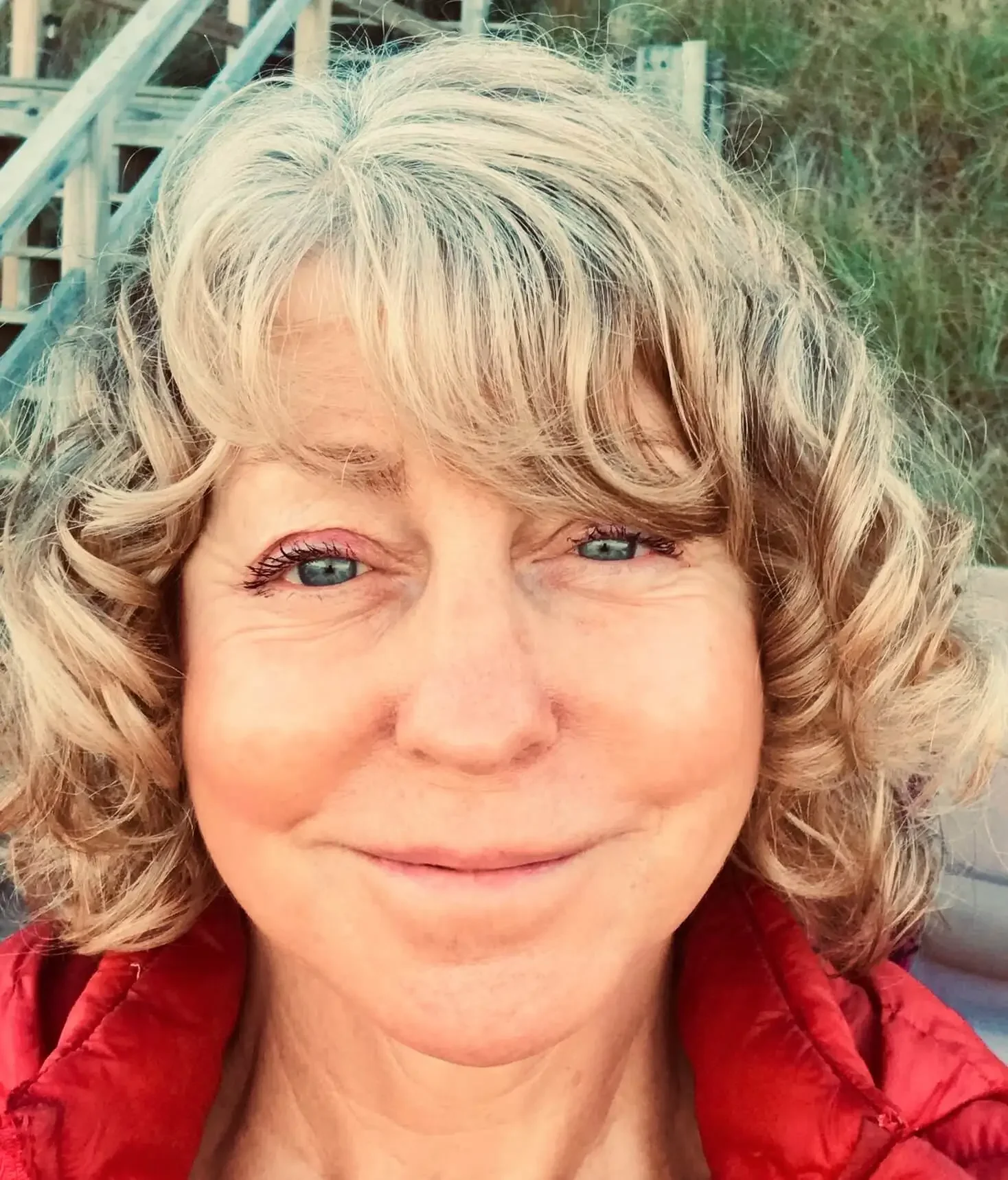 Close-up of a smiling woman with curly blonde hair and blue eyes wearing a red jacket outdoors.