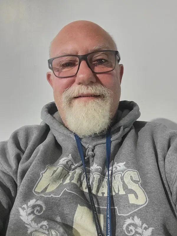 Close-up of a smiling older man wearing glasses and a gray sweatshirt with the word 'WILLIAMS' printed on it. He has a bald head and a white beard and mustache.