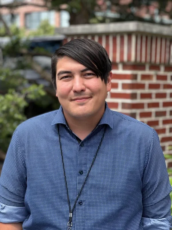 A young man with dark hair and a light complexion standing outdoors in front of a red brick building and greenery, smiling at the camera, wearing a blue button-up shirt and a lanyard.