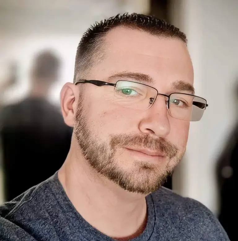 A man with short hair, glasses, and a beard, smiling at the camera in an indoor setting.