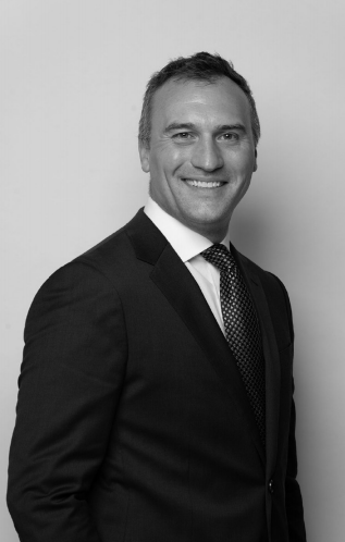 A hotel real estate professional smiling in a suit and tie photographed against a plain background.
