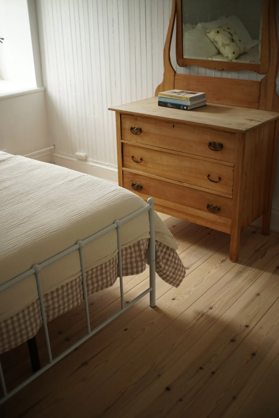 A bedroom corner with a white metal bed frame, a cream-colored quilt, and a wooden dresser with a mirror and books, adjacent to a window with vertical blinds, and a bed with a pillow reflected in the mirror.