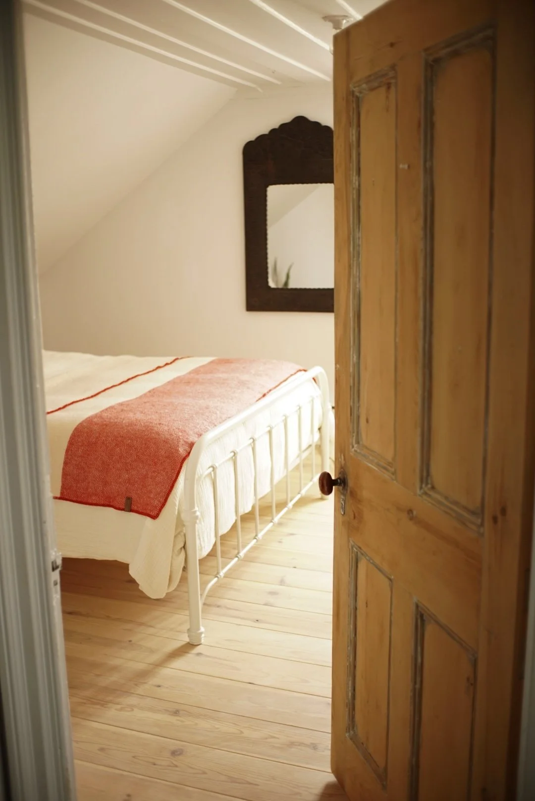 A cozy bedroom with a white metal bed frame, a red and white blanket, a wooden floor, a decorative mirror on the wall, and an open rustic wooden door.
