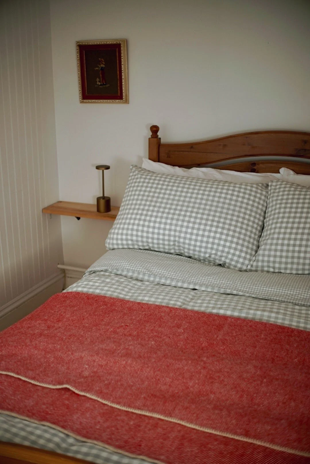 A neatly made bed with gingham checkered pillows and a red blanket in a bedroom with a wooden headboard, a small wall-mounted shelf with a lamp, and a framed picture hanging on a white wall.