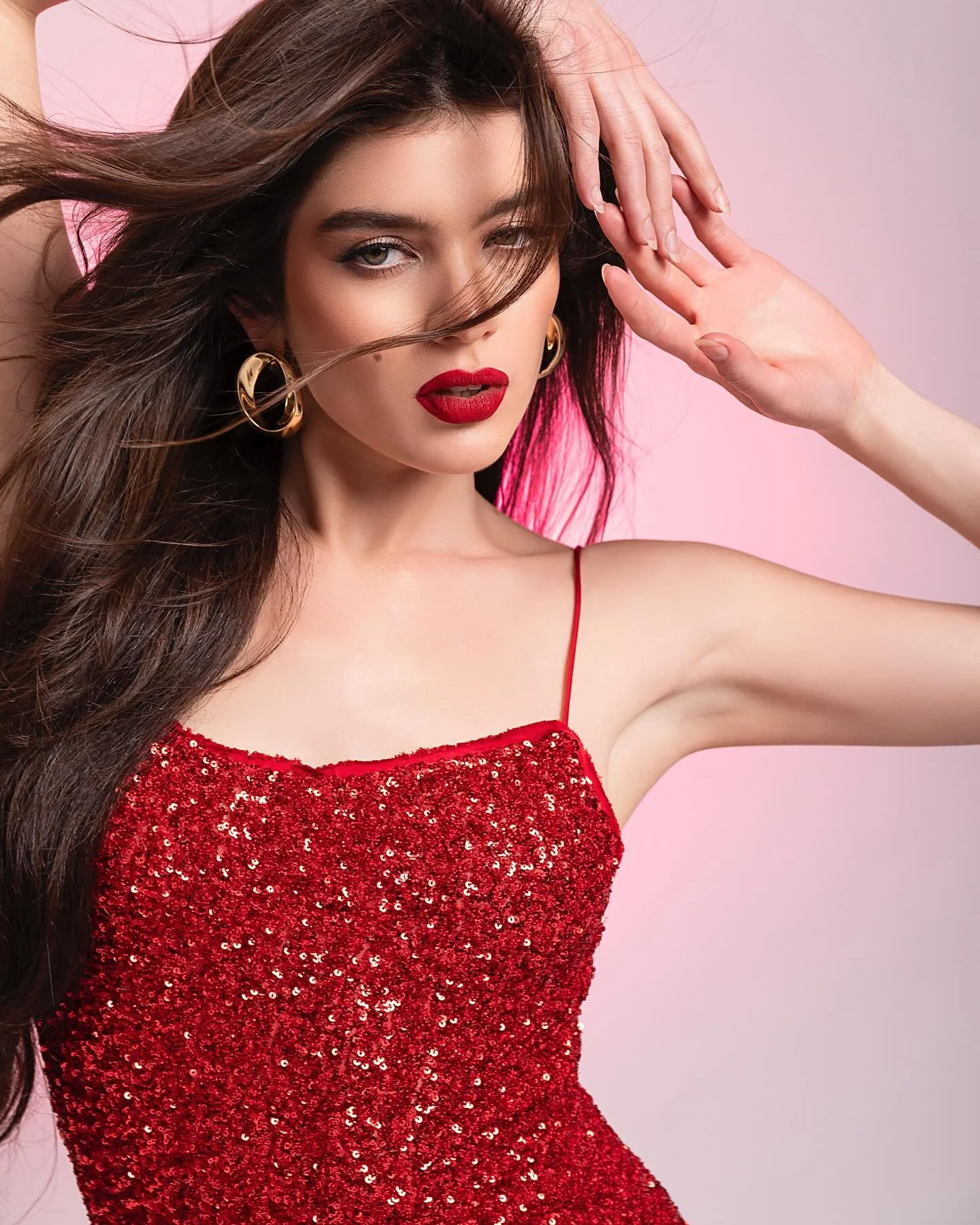 Mujer con cabello castaño largo y rizado, vestido rojo con lentejuelas, labios rojos y aretes dorados, posando con mano en la cabeza en fondo rosa.