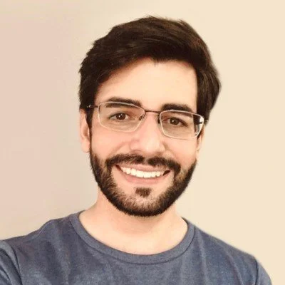 A smiling man with dark hair and glasses, wearing a bluish-gray shirt, against a light-colored background.