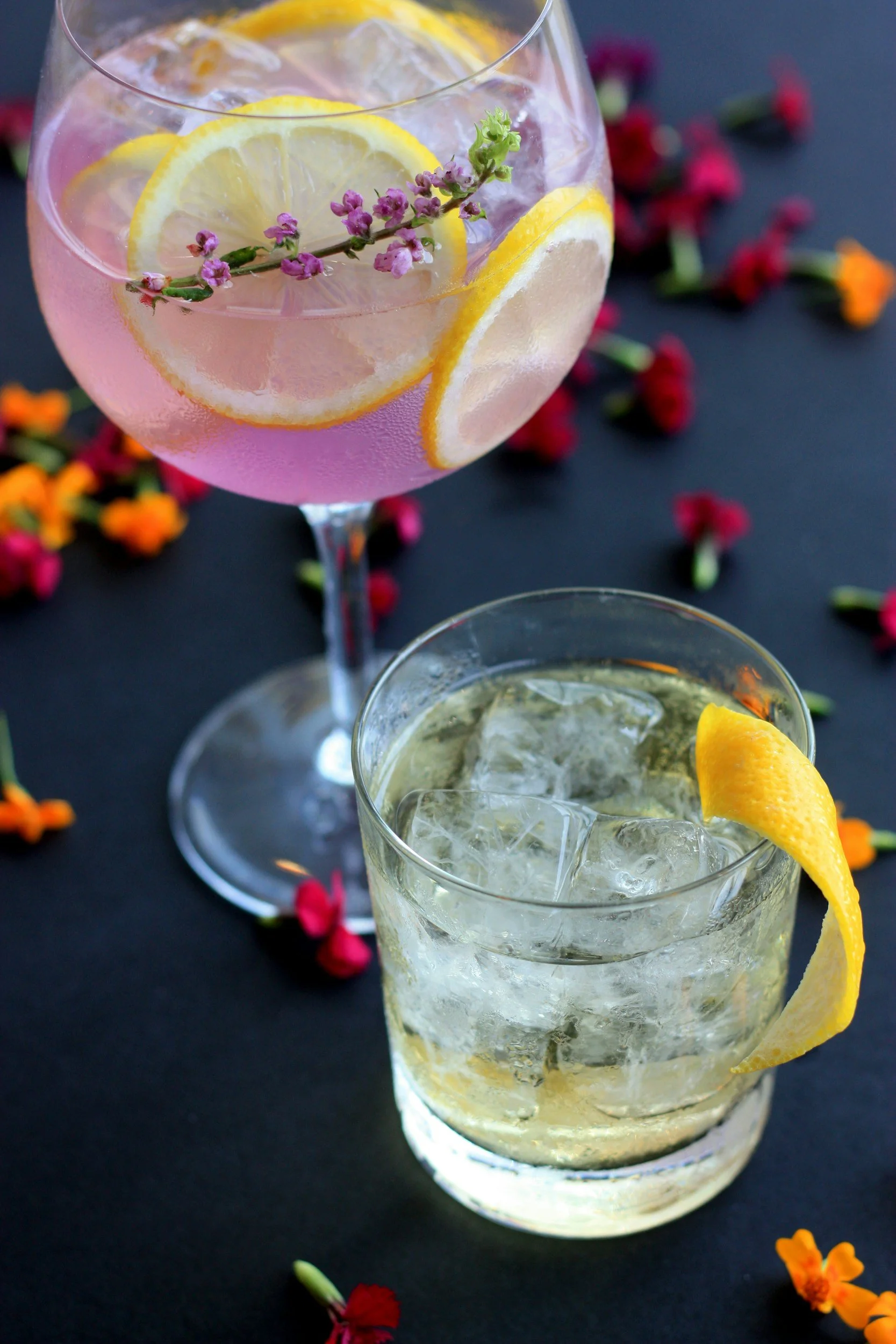 A glass of lemonade garnished with lemon slices and a sprig of purple flowers, with ice cubes inside. In the background, another drink with lemon and ice, surrounded by scattered small flowers on a dark surface.