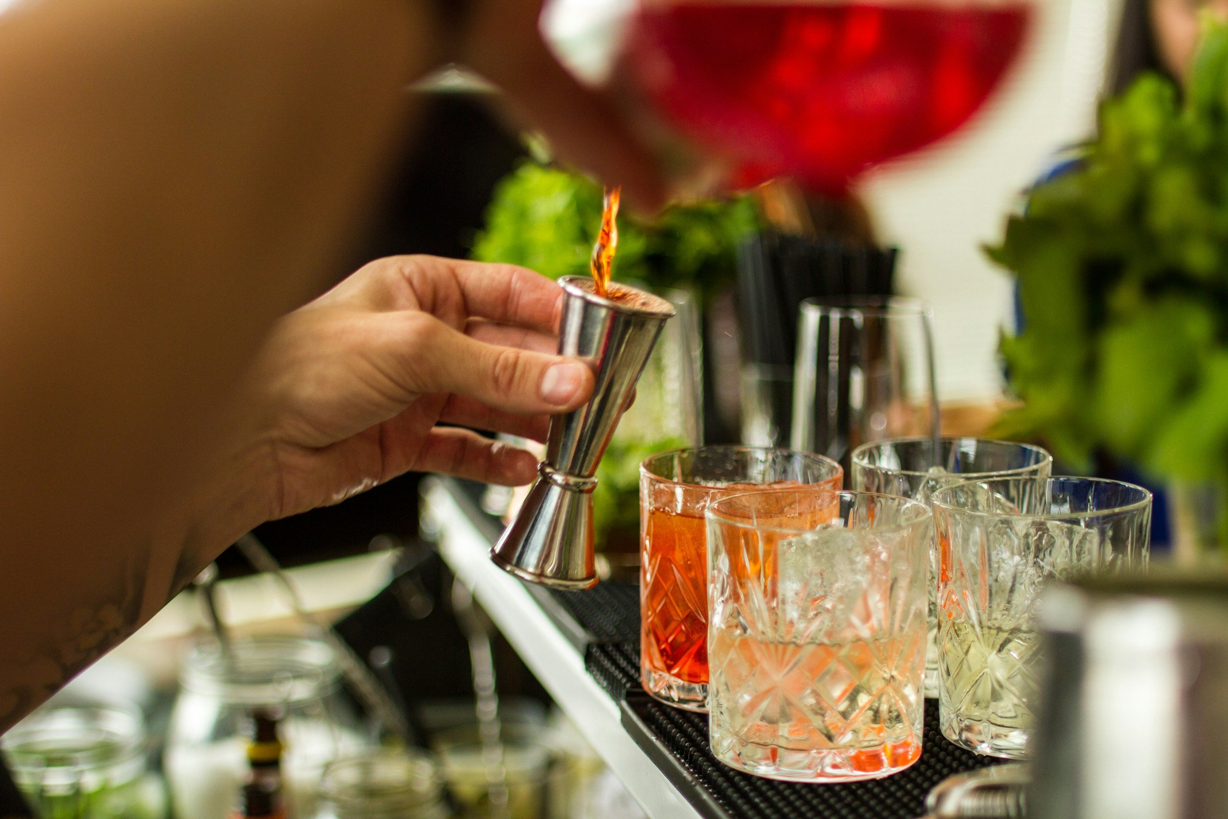A hand pouring a red drink from a small metal pitcher into a glass, with other glasses and green plants in the background.