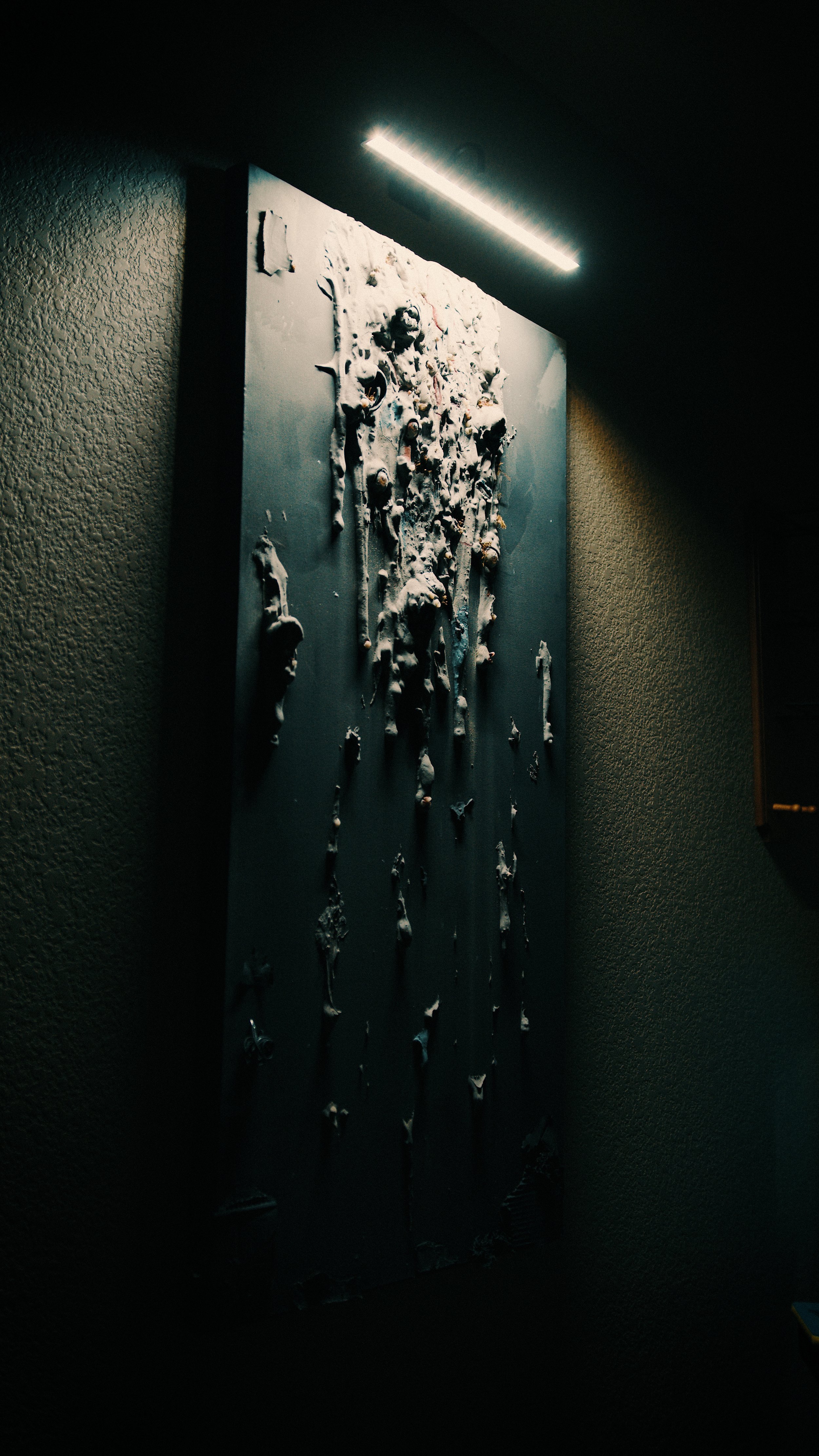 A dark, textured wall with illuminated artwork featuring textured, abstract shapes and figures on a vertical panel.