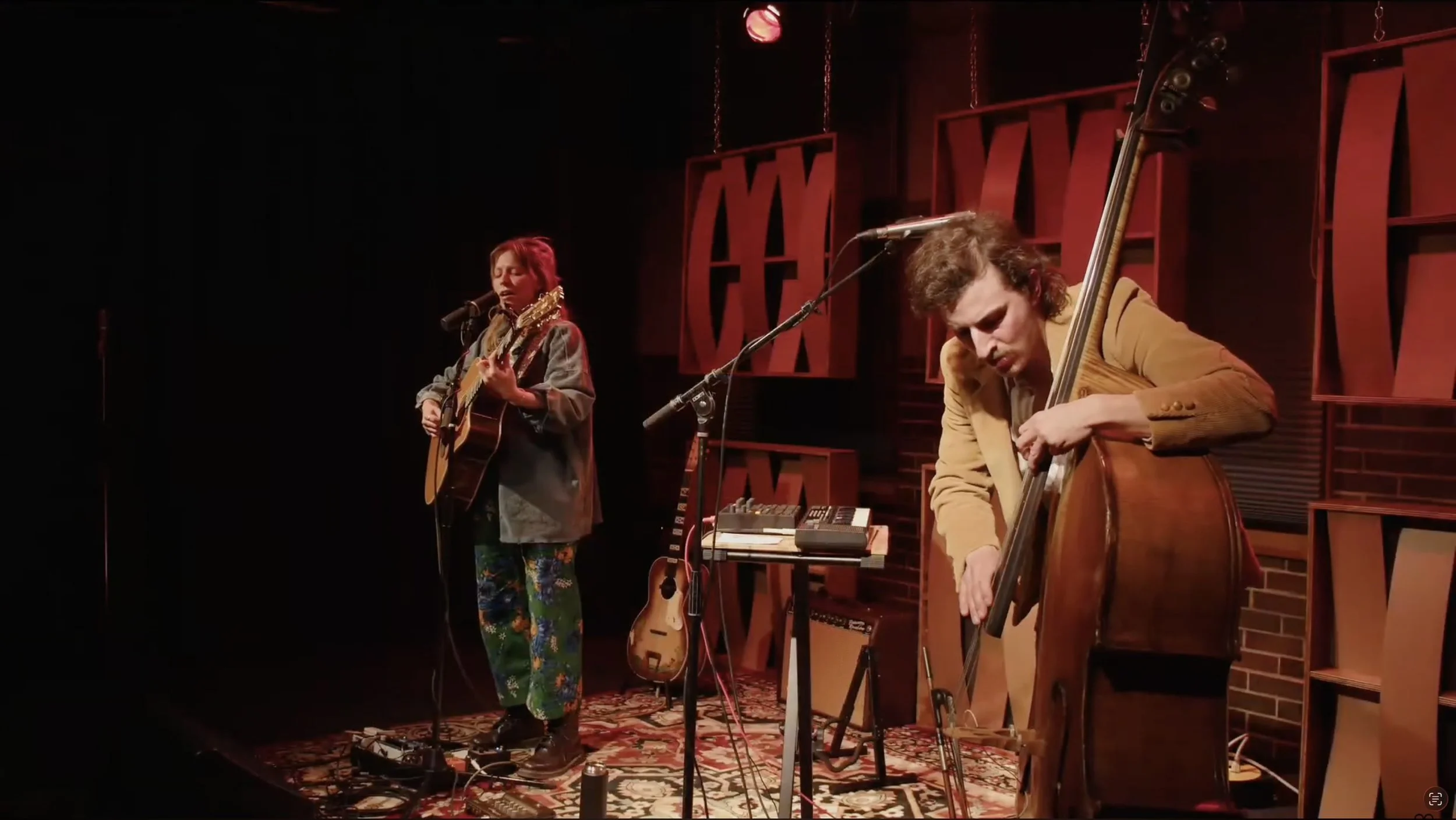 Two musicians performing on stage, one singing and playing guitar, the other playing upright bass, with various musical equipment around them.