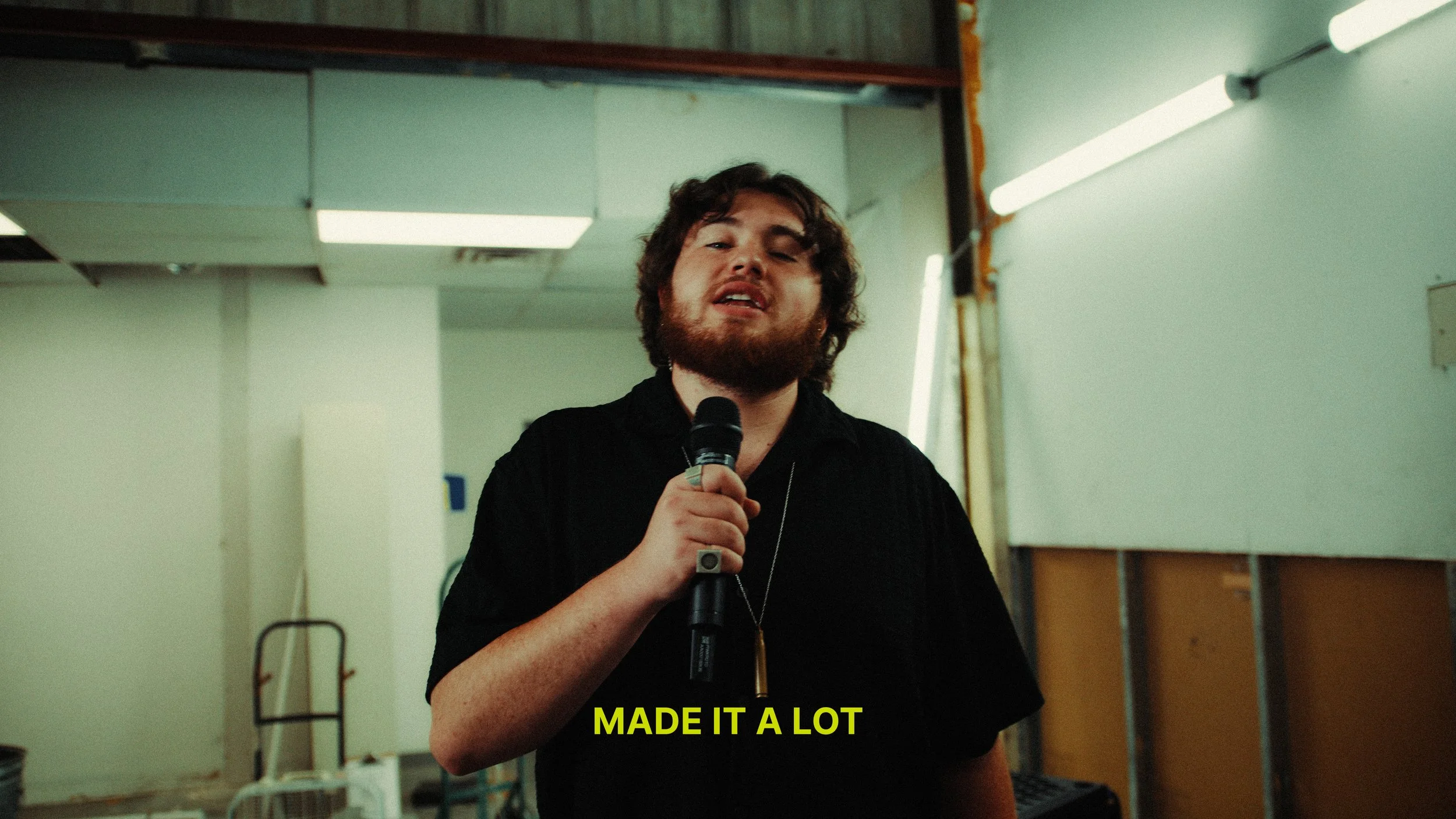 A man with curly brown hair and a beard is holding a microphone and speaking. He is wearing a black shirt and seems to be in a room with unfinished or industrial decor, including exposed drywall, fluorescent lights, and some equipment or furniture in the background. The image has a yellow caption at the bottom that reads, "MADE IT A LOT."