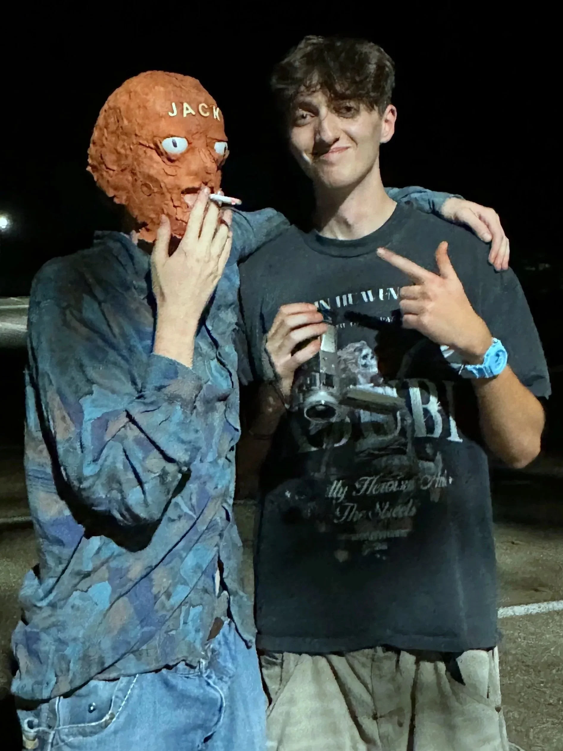 Two young men standing outside at night, one wearing a Jack Skellington mask with a cigarette, the other pointing at him and smiling.