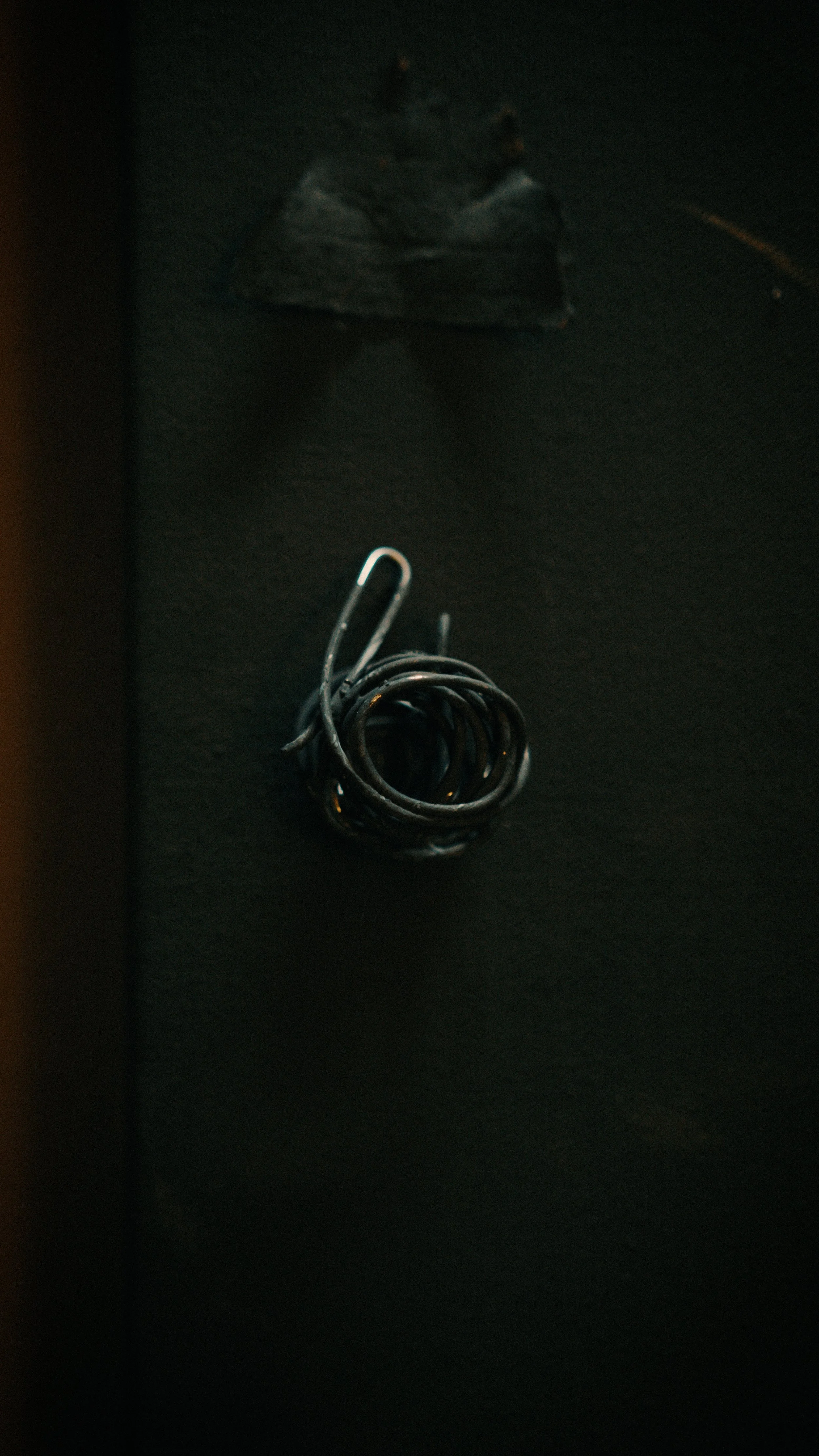 A black metal coil spring on a dark surface, with a small, blurry object behind it.
