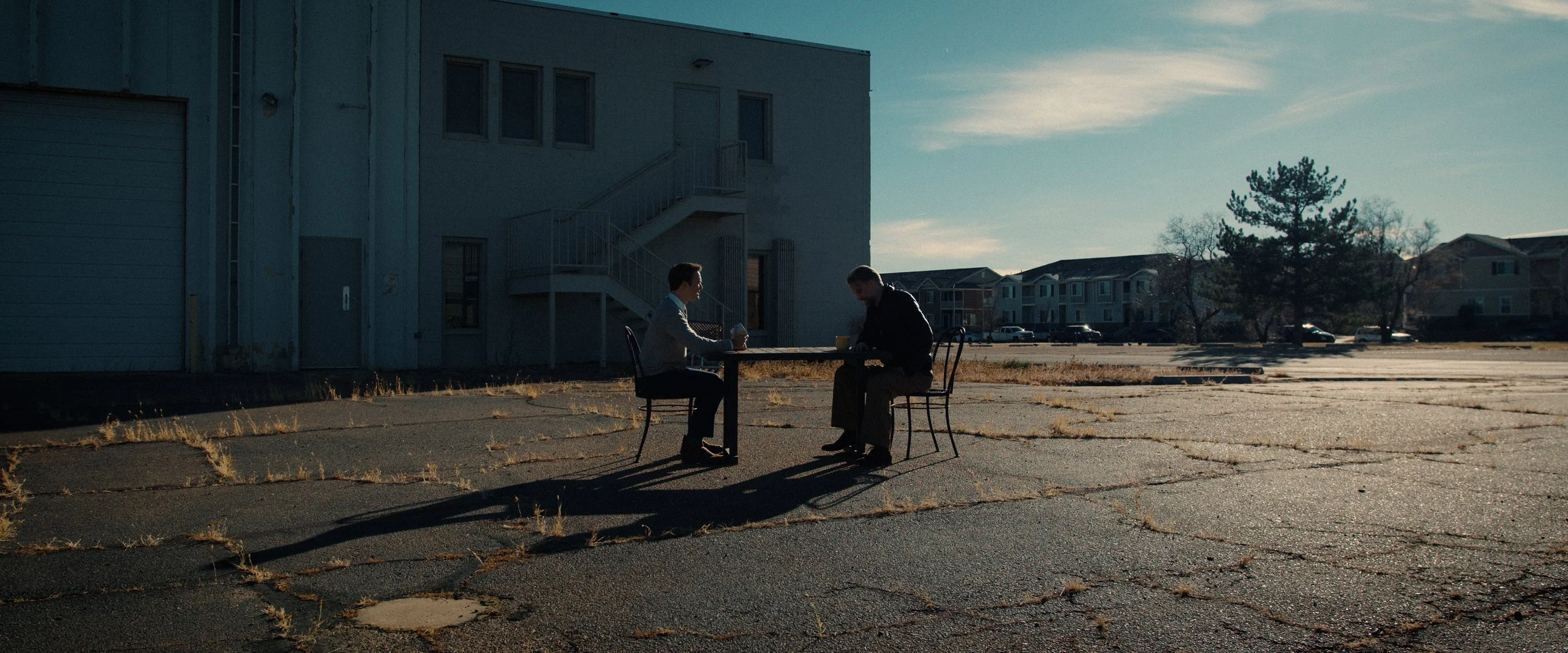 Two people sitting at a table outdoors near a building, sharing a meal on a sunny day, with a parking lot and trees in the background.