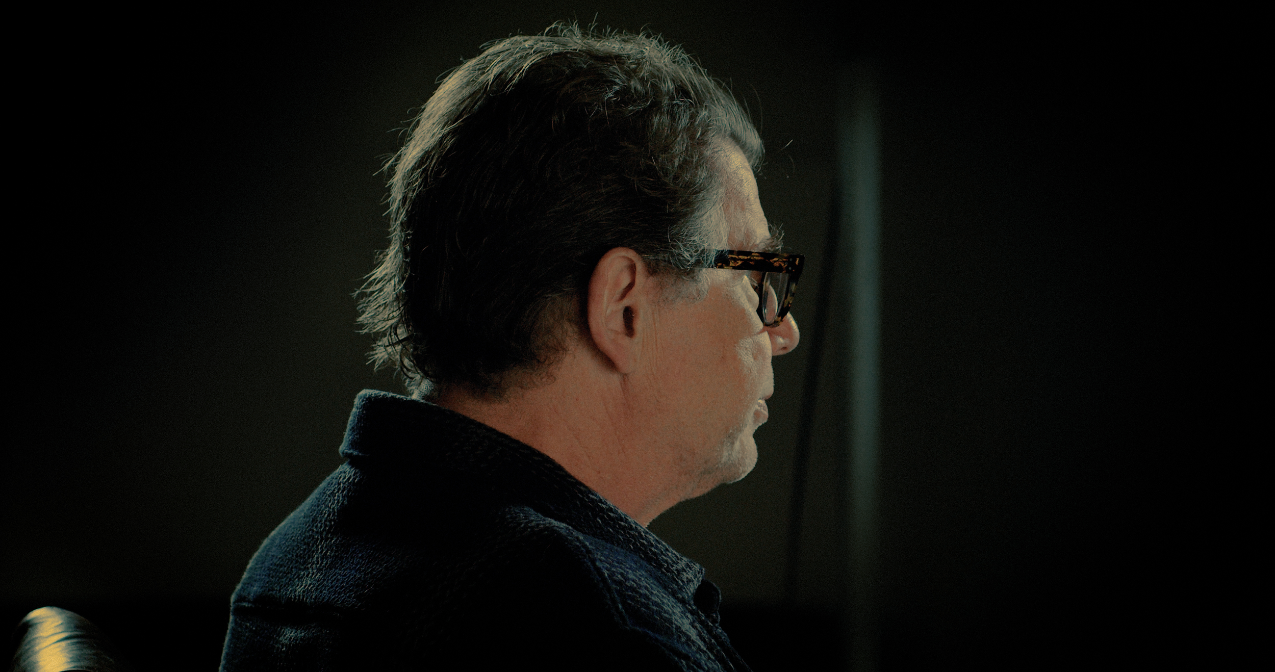 Side profile of a man with glasses and gray hair, sitting in a dimly lit room.