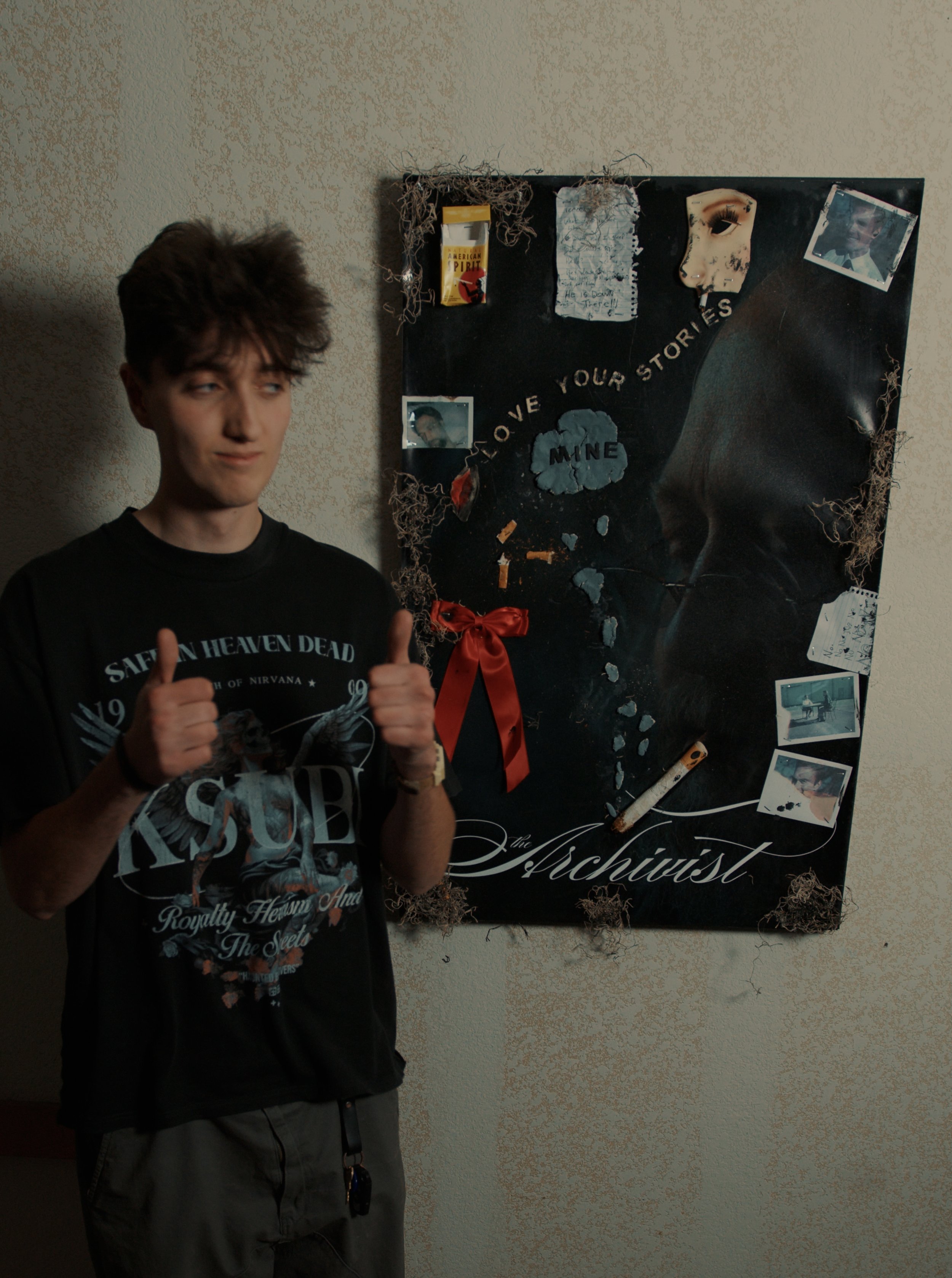 A young man with messy brown hair wearing a black Nirvana band t-shirt, holding up his thumbs, standing in front of a black poster with a mask, photos, and miscellaneous items, on a textured beige wall.