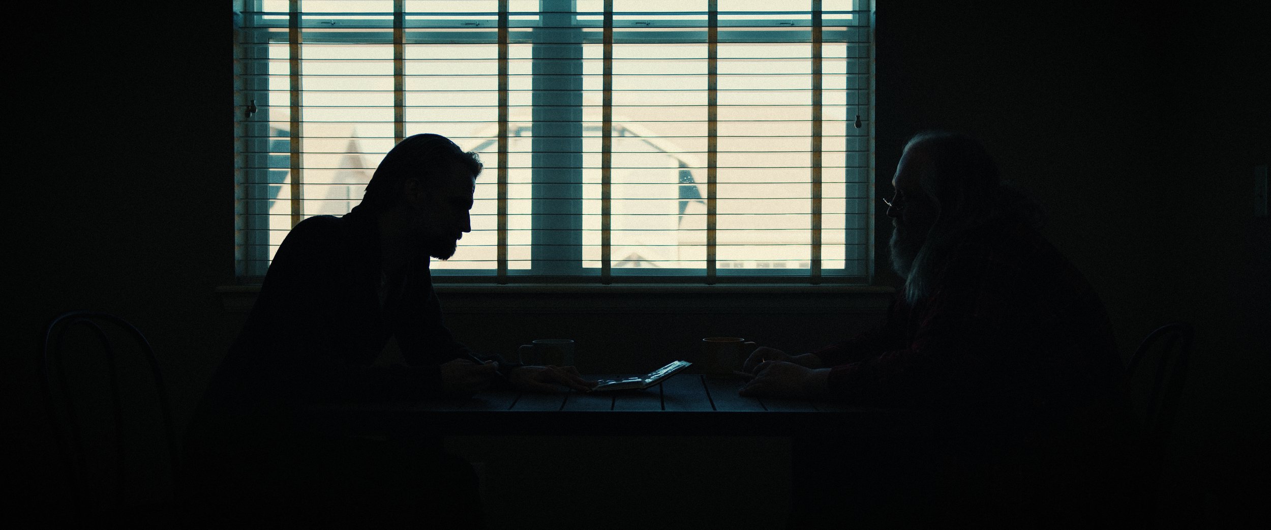 Two people sit at a table in a dark room, facing each other in silhouette, with a window with blinds behind them letting in light.