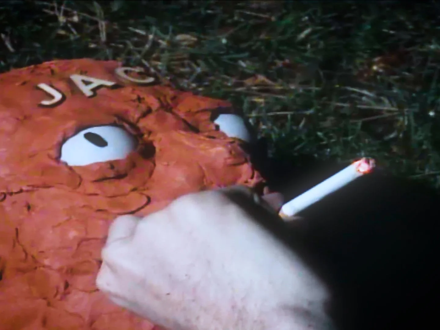A burnt cartoon-like face with large eyes and a cigarette in its mouth lying on the ground with grass nearby.