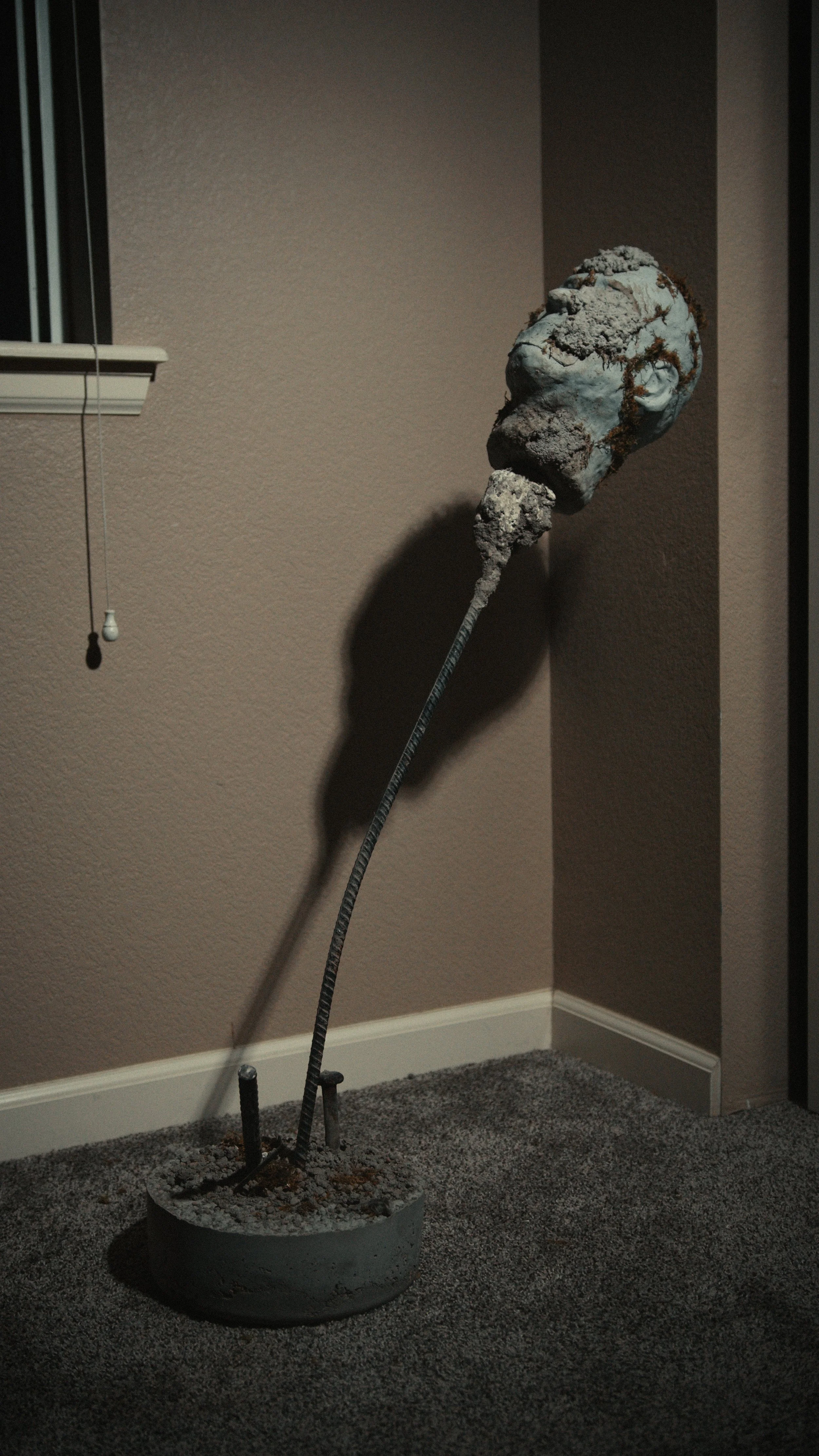 Interior corner of a room with a textured pink wall, small window with vertical blinds, and a potted plant-shaped sculpture made of concrete and metal standing on a gray carpet.