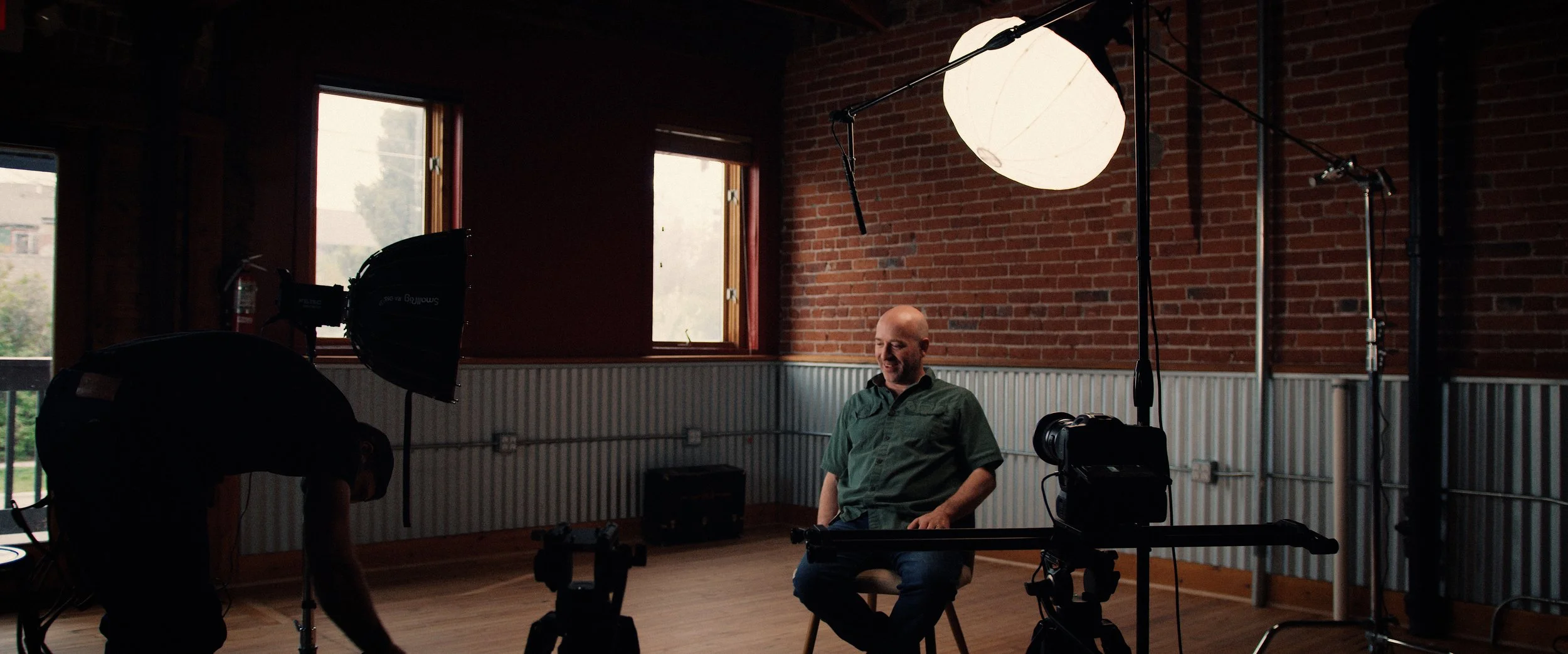 Man seated in a photography studio with professional lighting equipment, brick wall, and windows in the background.