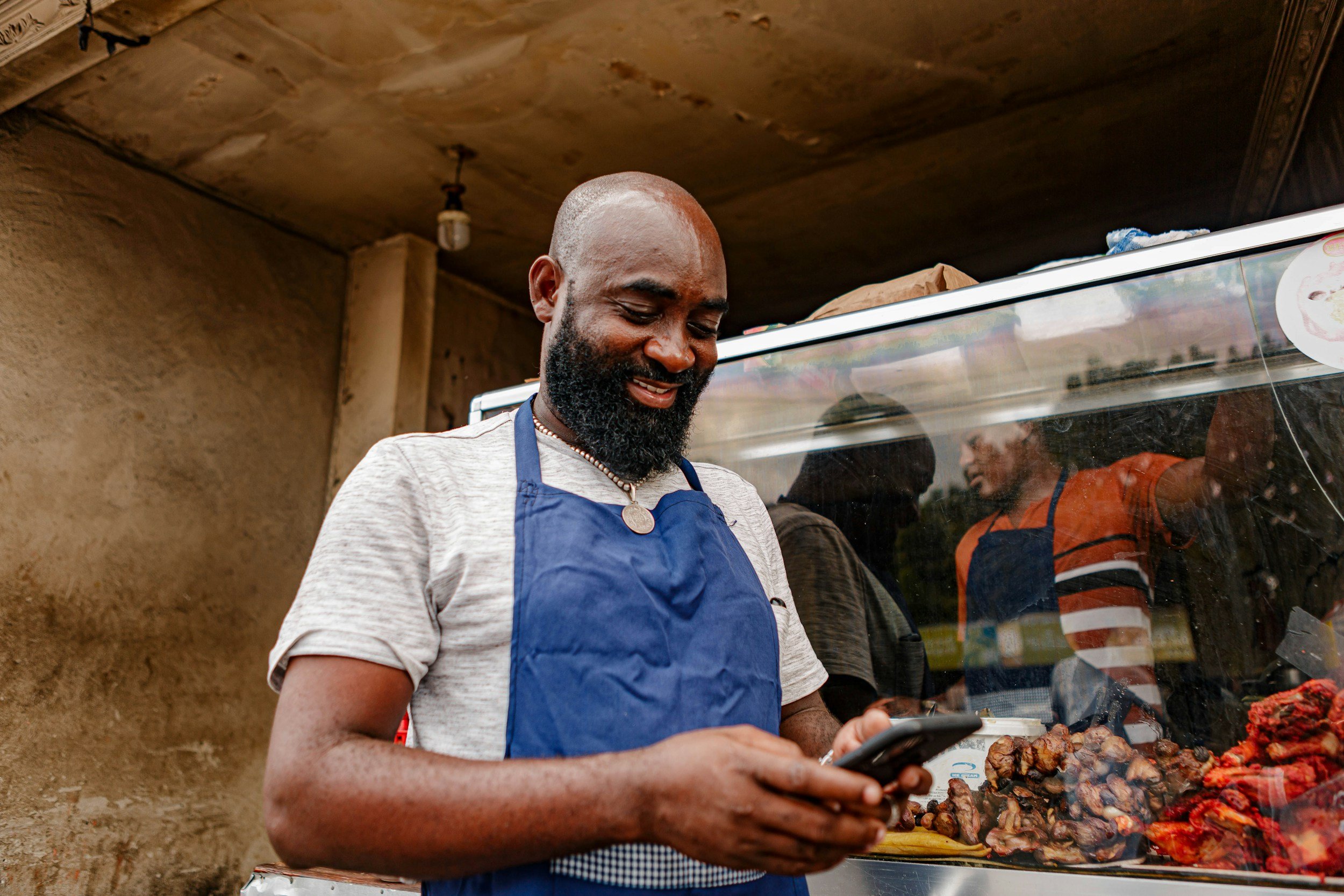 A man with a beard wearing a blue apron and a gray shirt, looking at his phone, standing behind a food stand with various meats on display, outdoors in front of a brown wall with a glass display case.