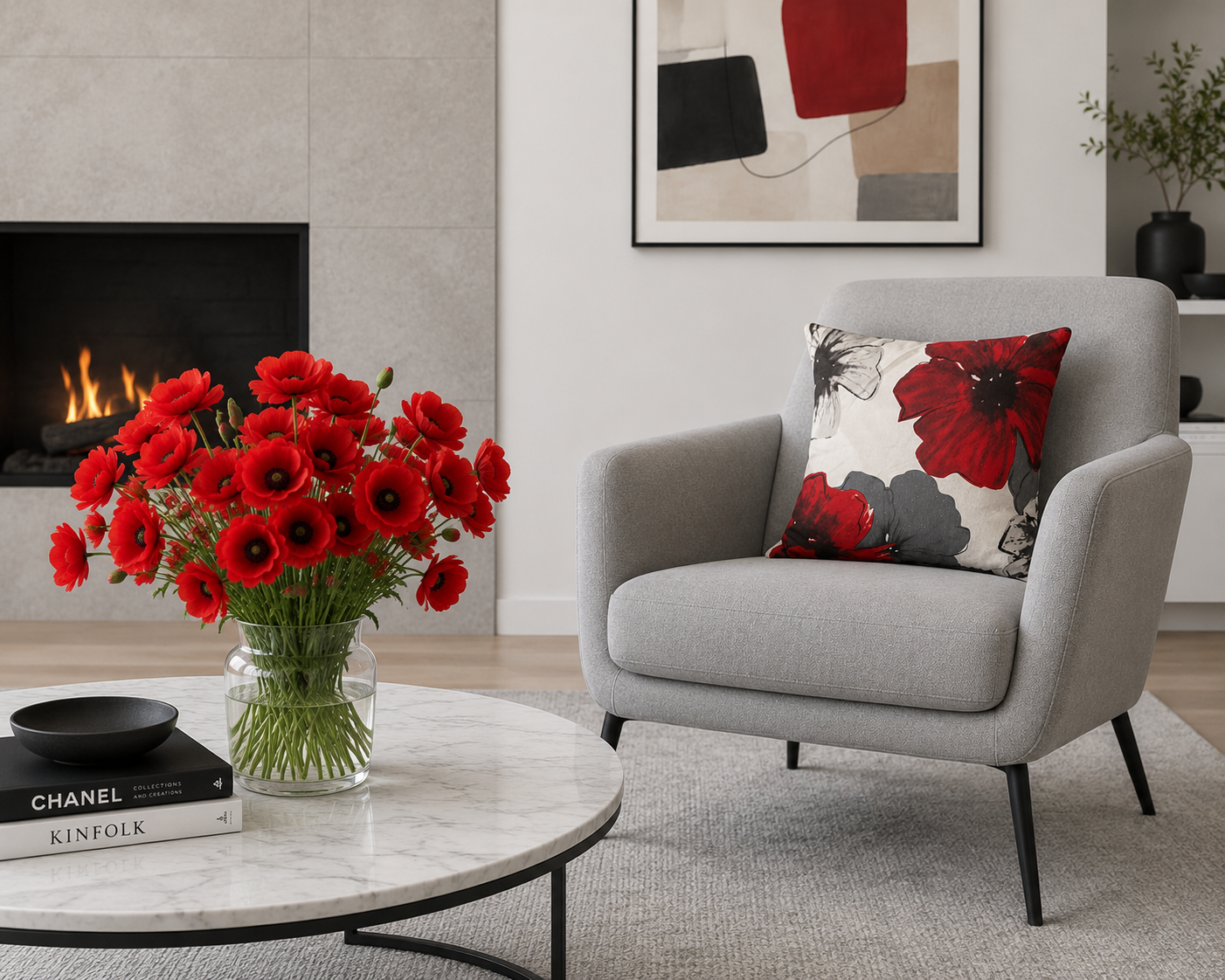 Living room with a gray armchair, a floral throw pillow, a round marble-top coffee table with a glass vase of red flowers, and a fireplace with a lit fire. There is a black bowl, two books, and artwork on the wall.