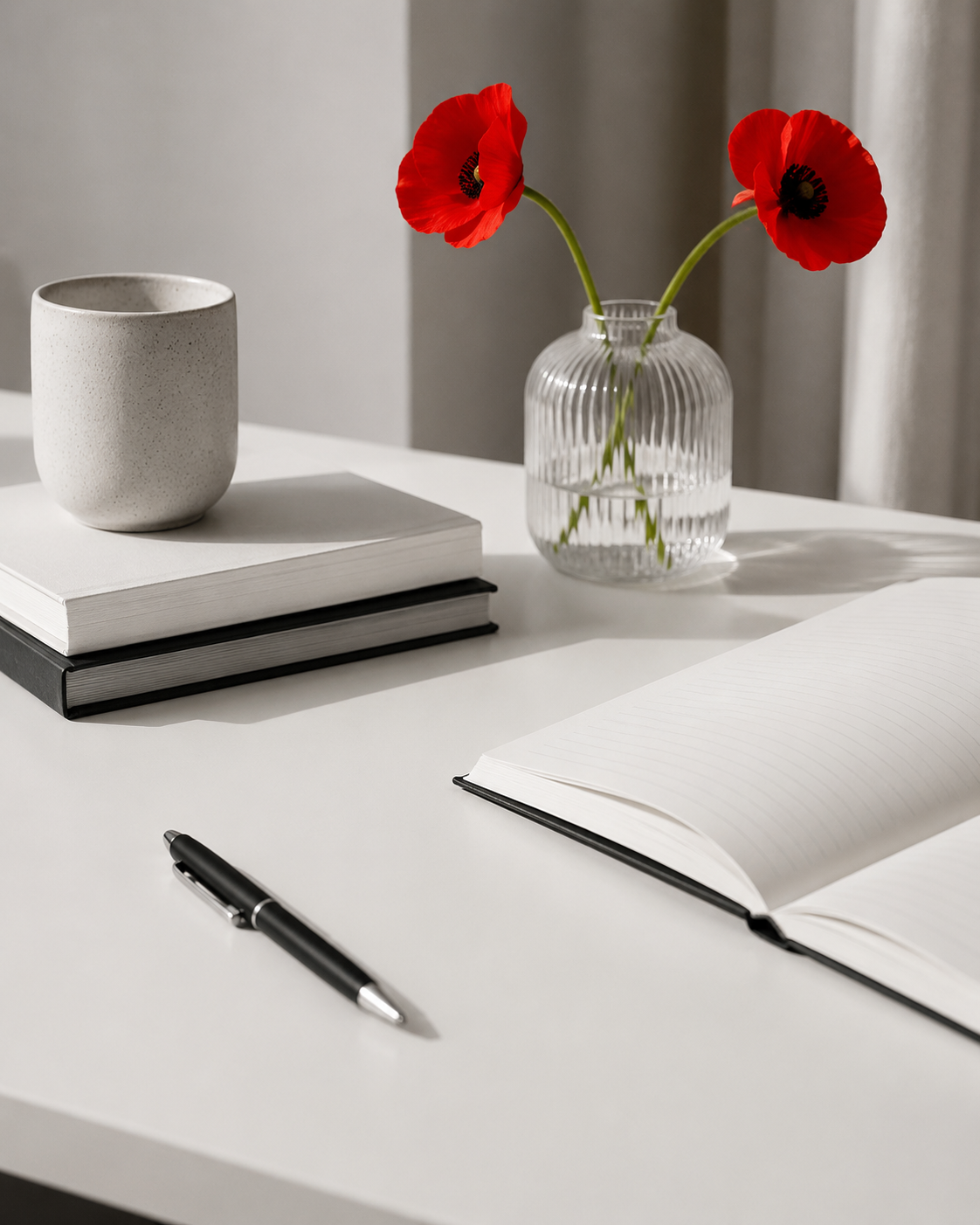 A white desk with a pen, an open notebook with lined pages, a closed black book, a white ceramic cup, and a glass vase with two red poppies.