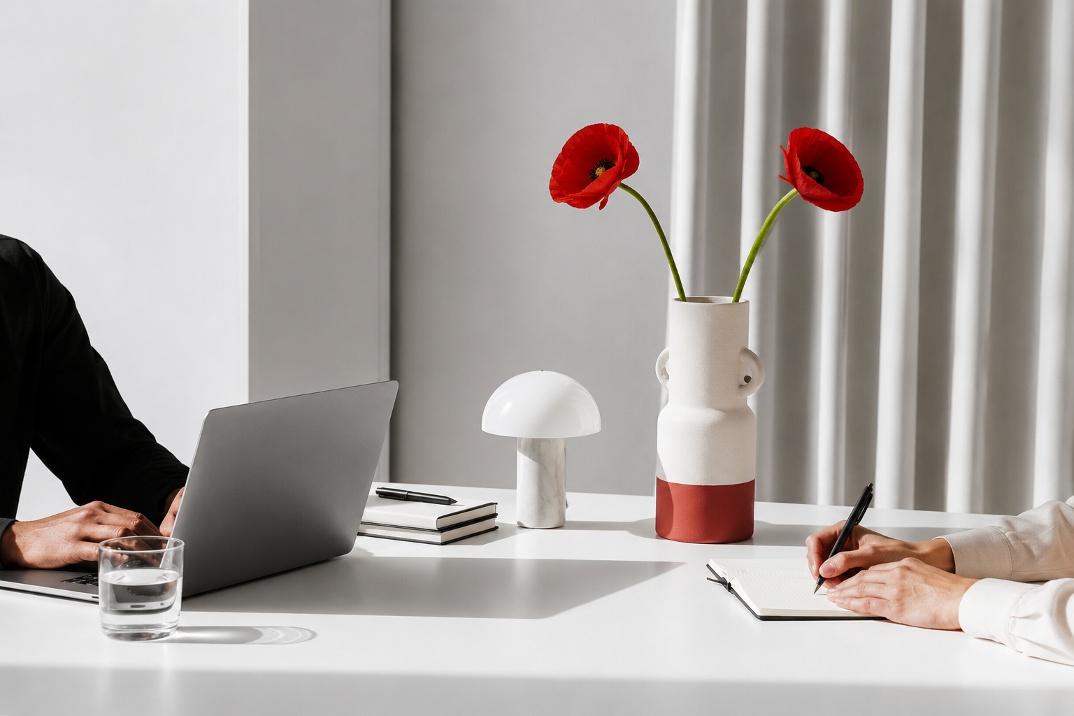 Modern office desk with a person's hand using a laptop, a glass of water, a small mushroom-shaped lamp, a white and red vase with two red poppies, and a person's hand writing in a notebook.
