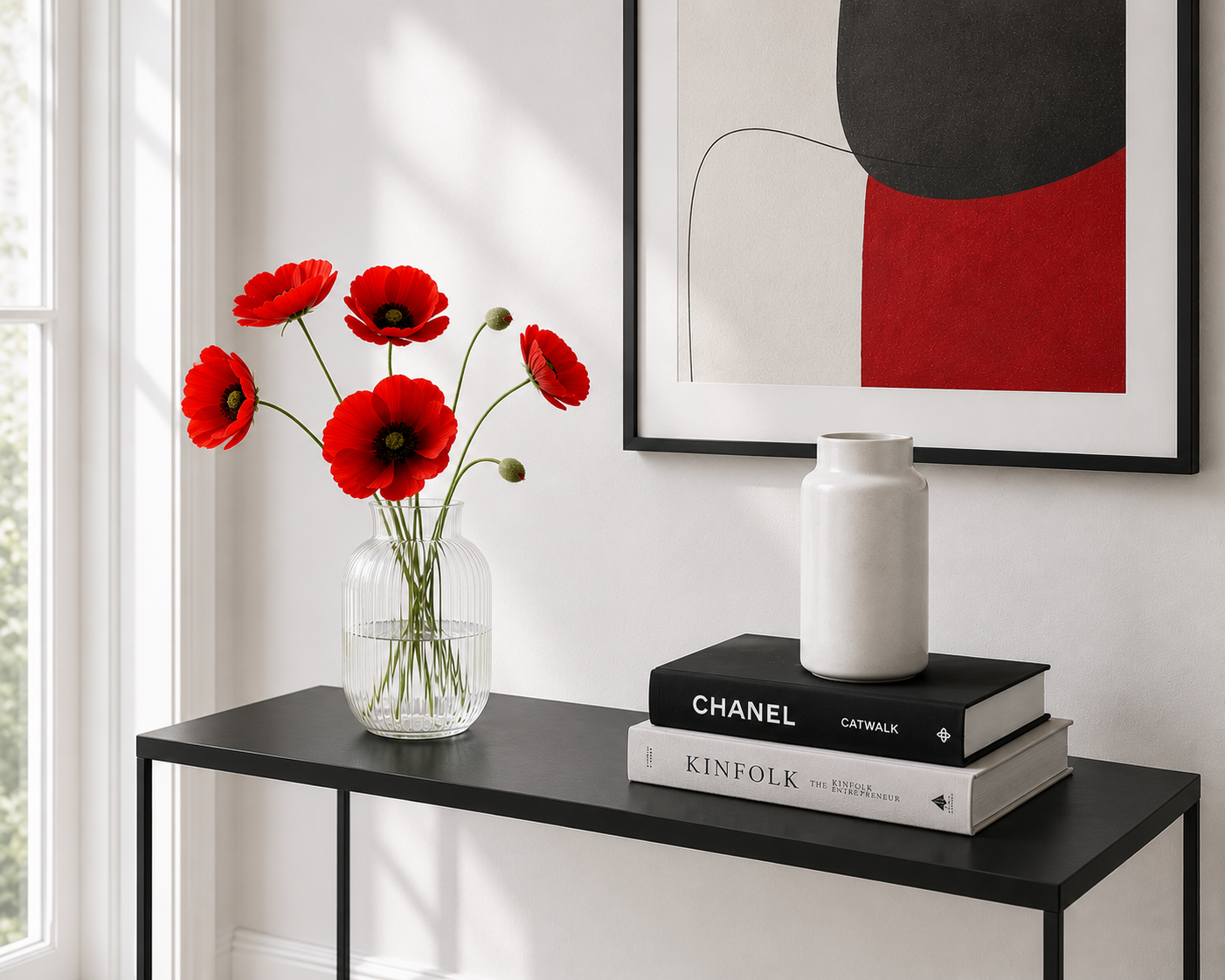 Red poppies arranged in a clear glass vase on a black console table beside stacked books and a white ceramic jar, with abstract artwork on a white wall in the background.