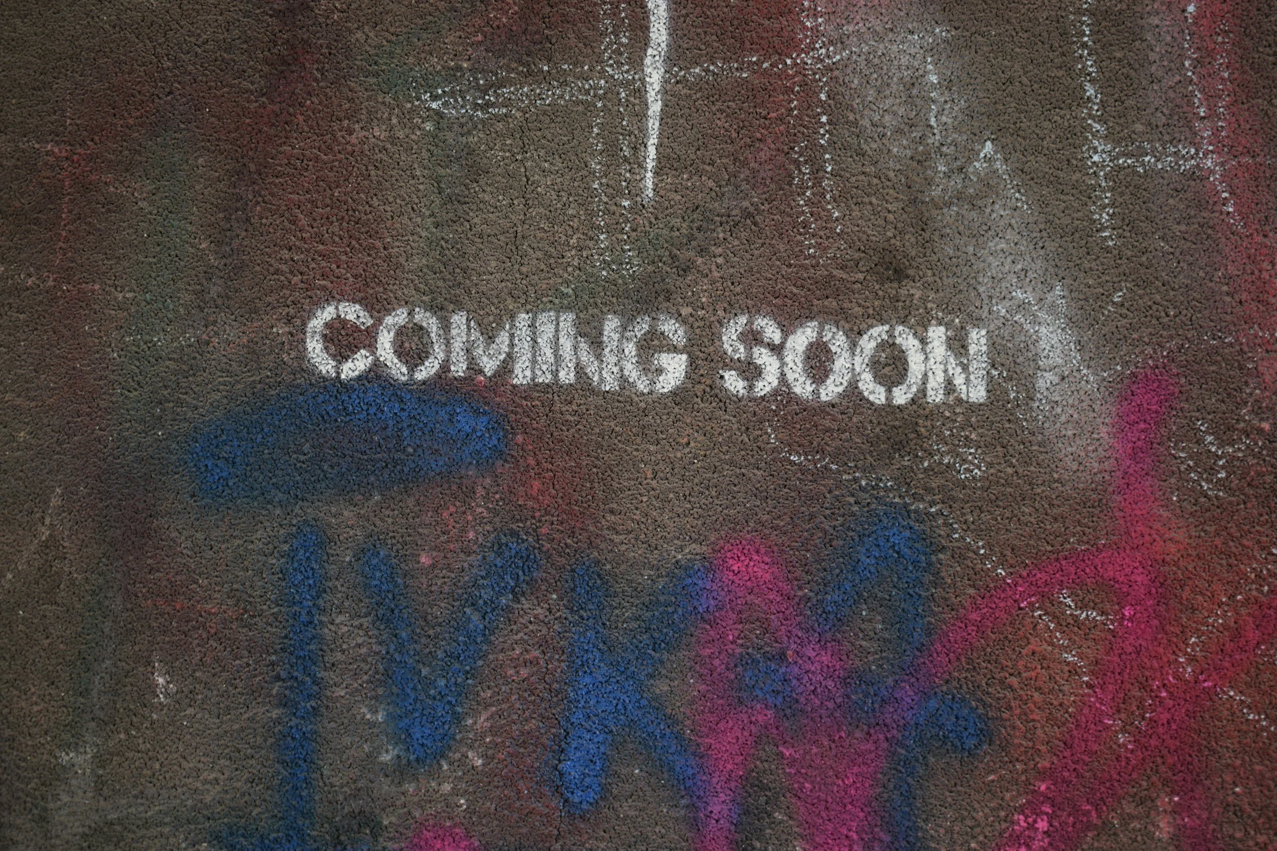 The words 'COMING SOON' written in white chalk on a gravel surface with colorful spray paint splatters around.