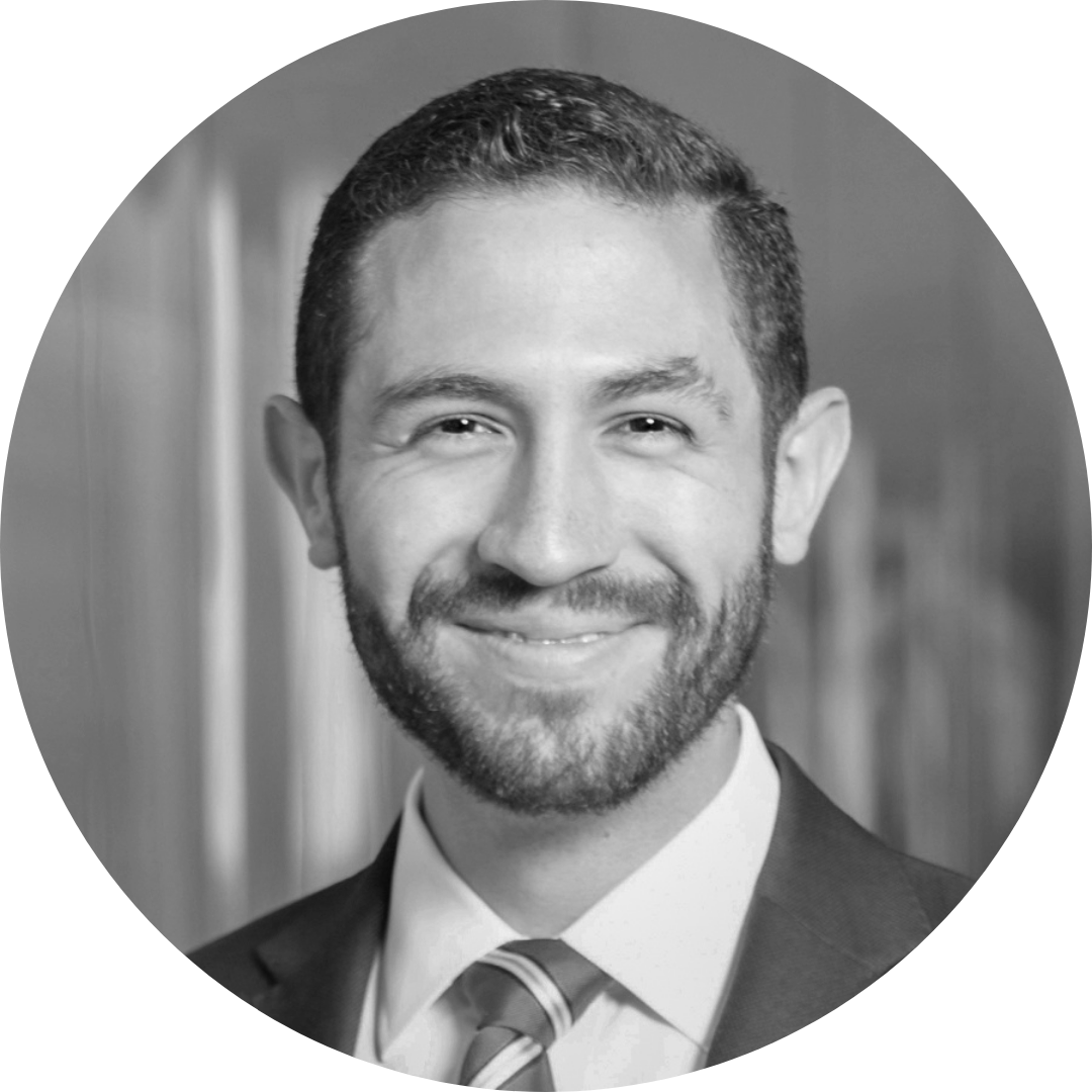 Black and white professional headshot of a smiling man with short hair, beard, wearing a suit and tie, in front of a blurred background.
