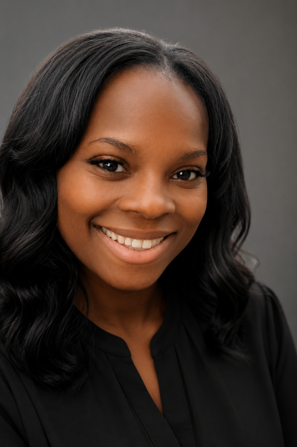 A young African American woman with long, wavy black hair smiling at the camera against a gray background.
