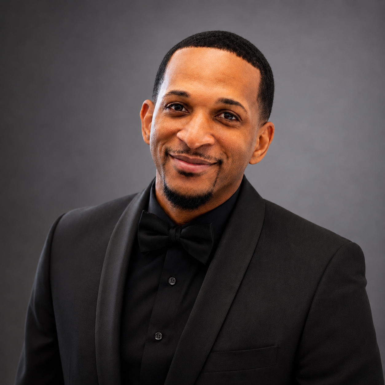 Headshot of a man in a black tuxedo with a bow tie, smiling at the camera, against a dark gray background.