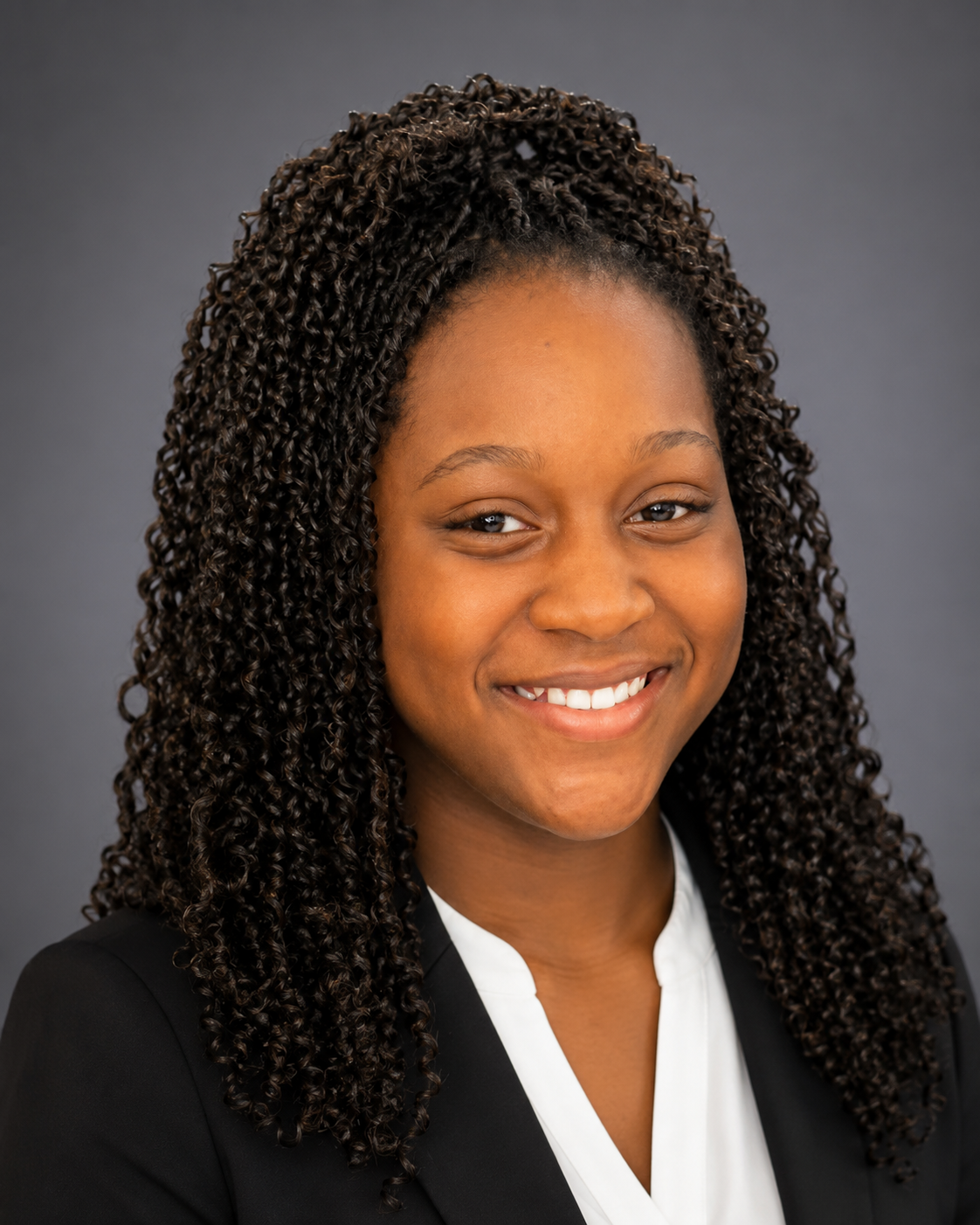 Headshot of a young African-American woman with curly hair wearing a black blazer and white blouse, smiling against a gray background.