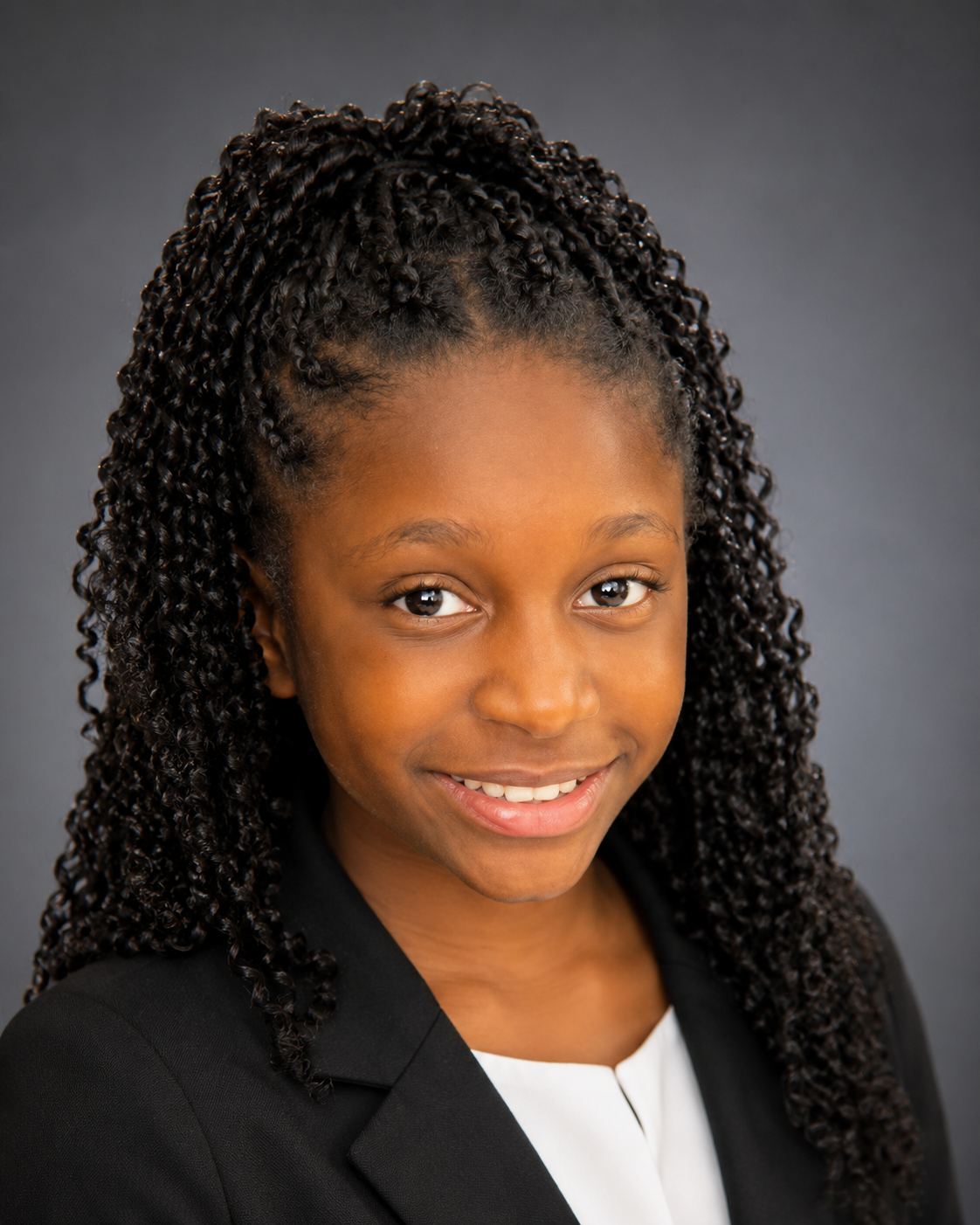 Headshot of a smiling young African American girl with curly hair, wearing a black blazer and white shirt, against a gray background.