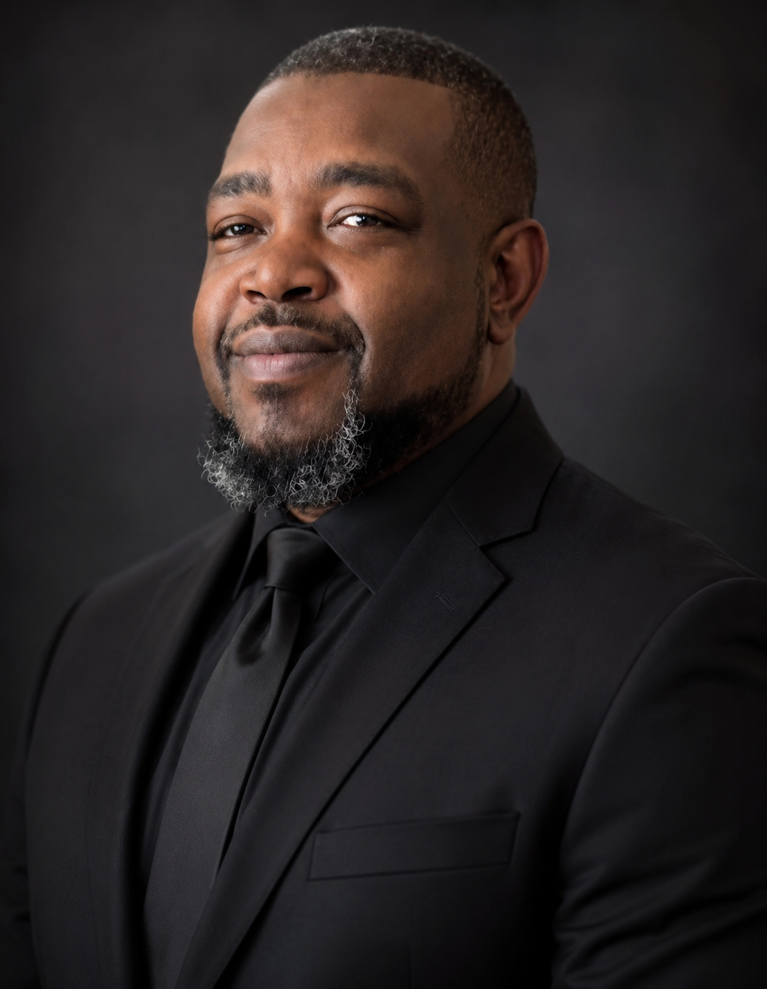 A confident African American man wearing a black suit and tie, with a short haircut and a beard with some gray, smiling subtly against a dark background.