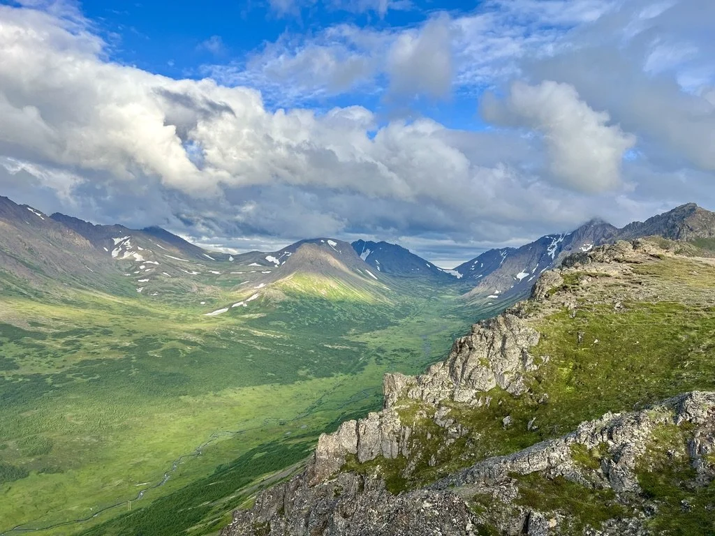 Scenic mountain landscape with green valleys, rugged cliffs, and snow patches on mountain peaks under a partly cloudy sky.
