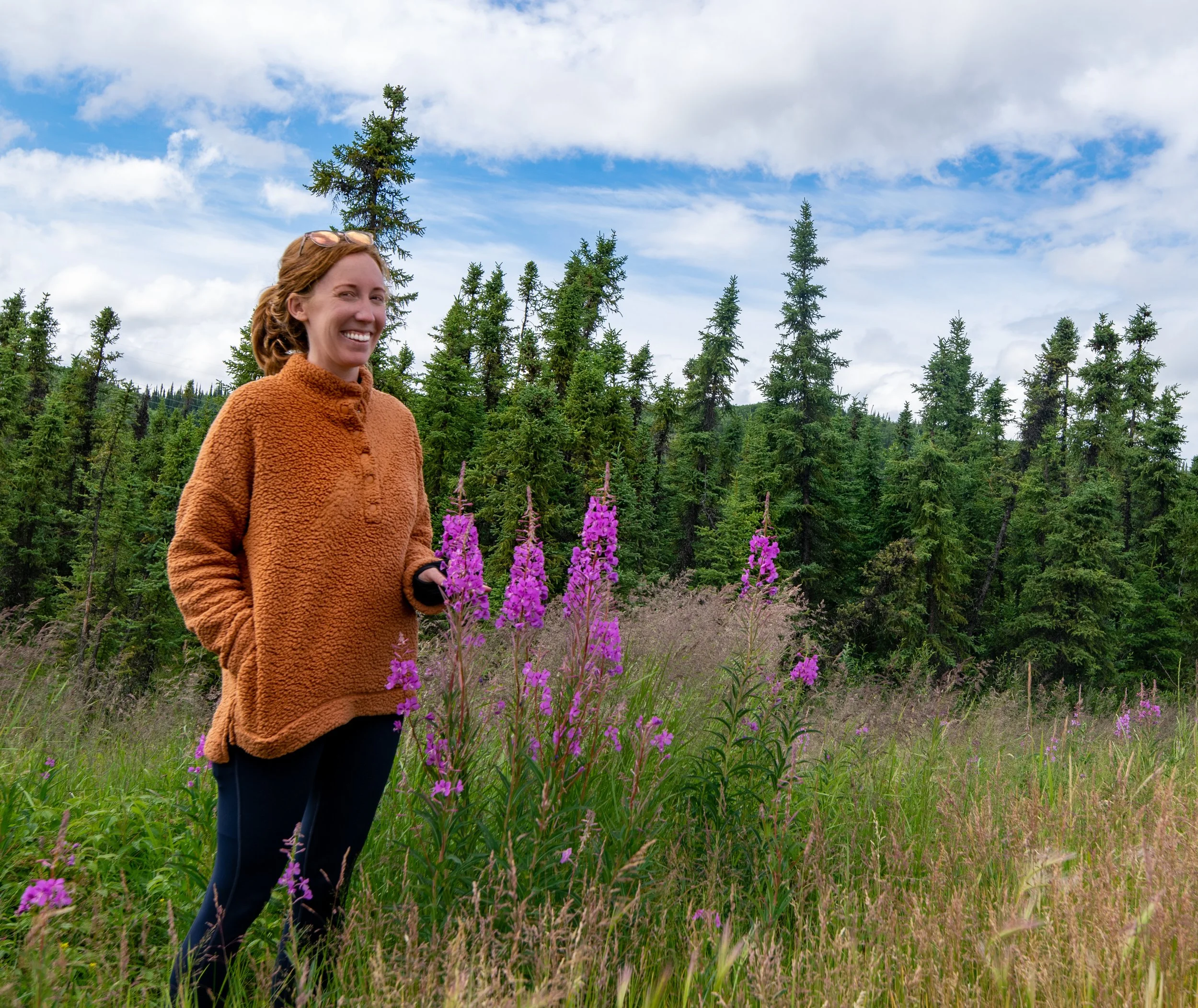 Woman smiling and holding pink flowers in a green meadow surrounded by trees under a partly cloudy sky.