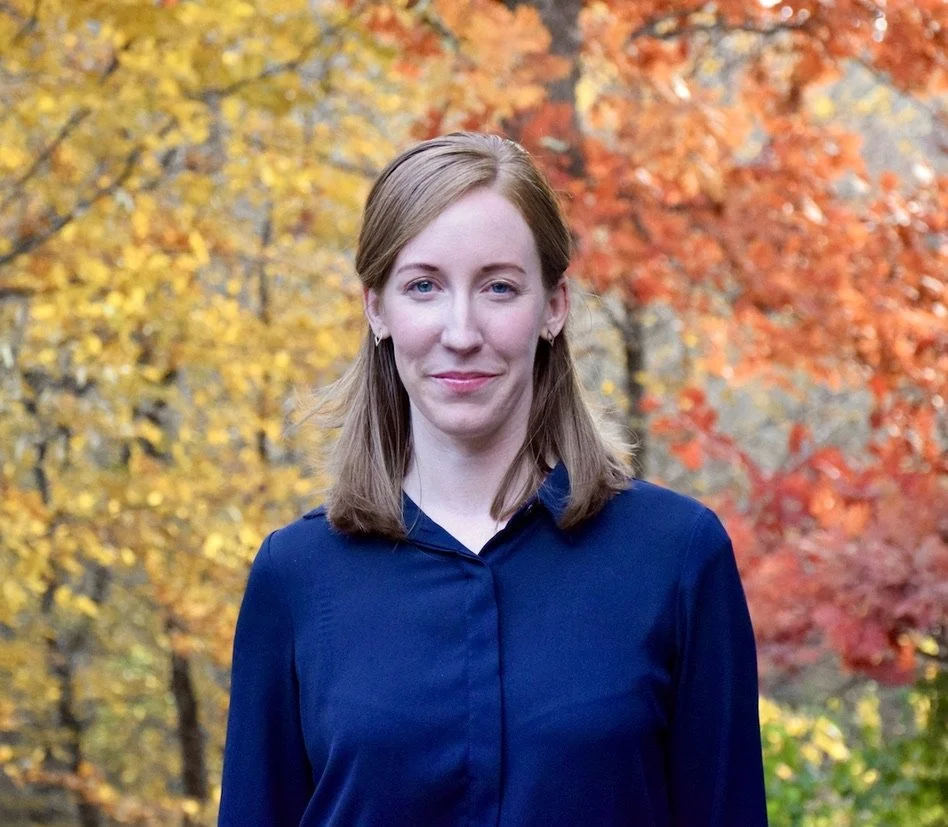 A young woman with shoulder-length light brown hair, wearing a dark blue shirt, standing outdoors among autumn-colored trees with yellow and red leaves.