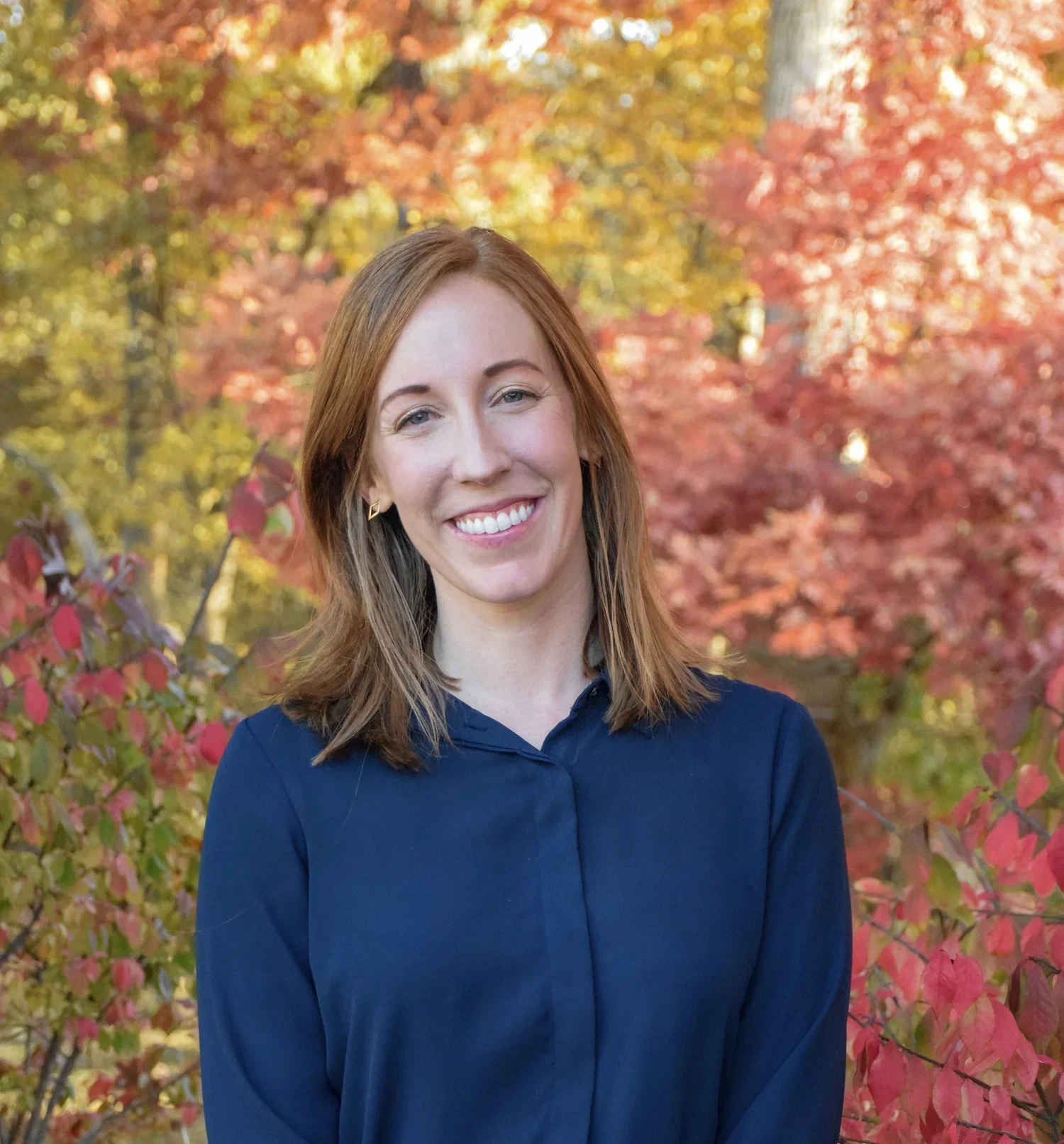 A woman with shoulder-length brown hair and blue eyes, smiling, wearing a navy blue shirt, standing outdoors in front of a background of colorful autumn trees with red, orange, yellow, and green leaves.