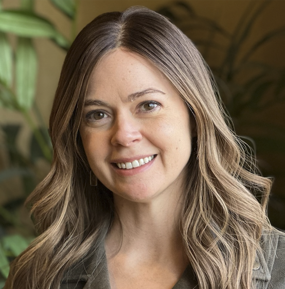 A woman with shoulder-length wavy hair smiling at the camera, background with green plants.
