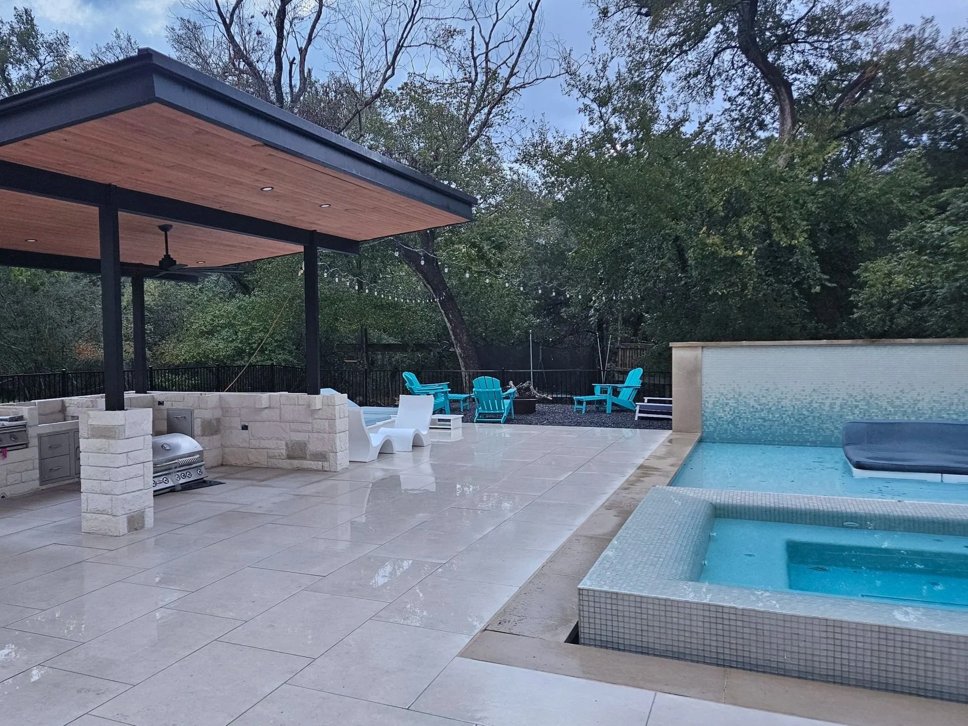 An outdoor patio with a swimming pool, blue chairs, a covered seating area with a ceiling fan, and string lights. Trees surround the area.