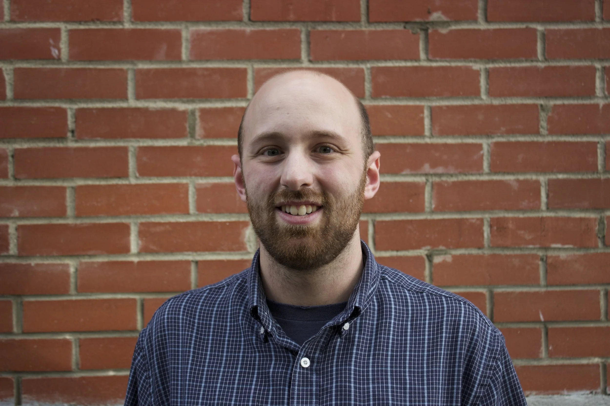 A smiling man with a beard and short hair standing in front of a red brick wall, wearing a blue checked shirt.