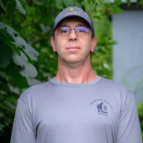Man wearing glasses, a baseball cap with 'GNOME' logo, and a light gray shirt with 'Vancouver Aquarium Gnome' logo, standing outdoors with green foliage in the background.