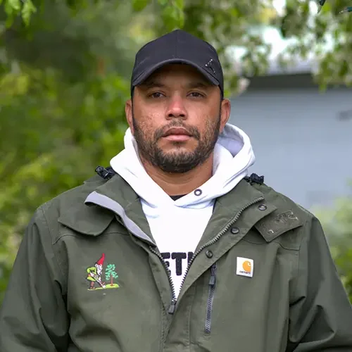 Man wearing a black cap, white hoodie, and green Carhartt jacket outdoors with trees in the background.